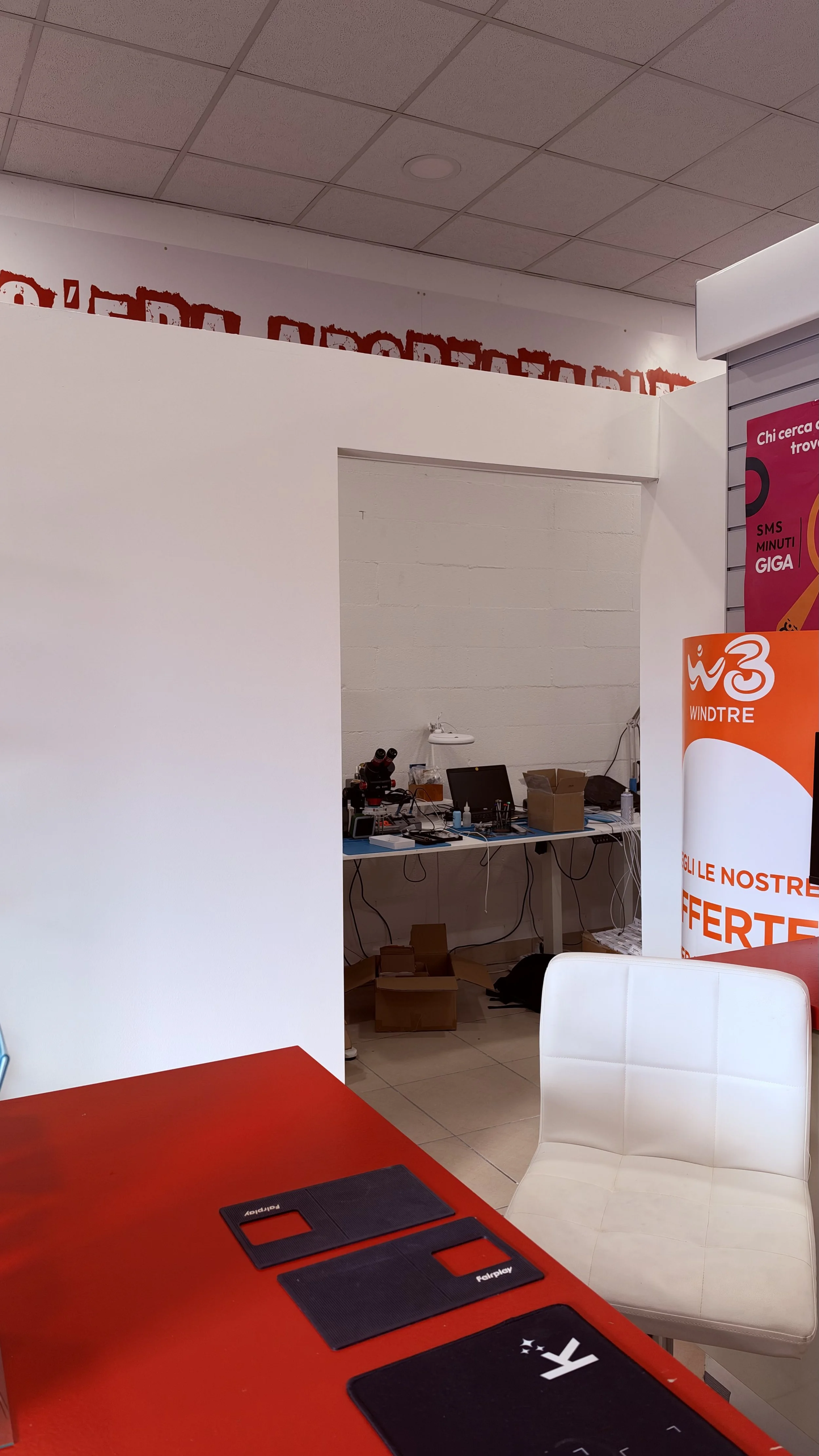 Interno di un ufficio o spazio di lavoro con tavoli, sedie e attrezzature tecnologiche. Parete con poster pubblicitari e scaffali vuoti o poco pieni.