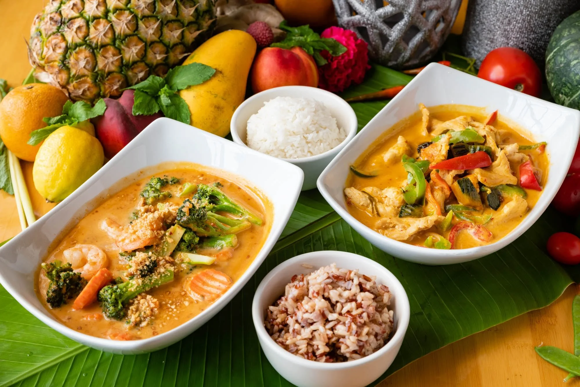 Assorted Asian dishes including curry with vegetables and shrimp, served with white rice and brown rice, surrounded by fresh fruits such as pineapple, oranges, apple, lemon, and tomatoes on a bamboo leaf background.