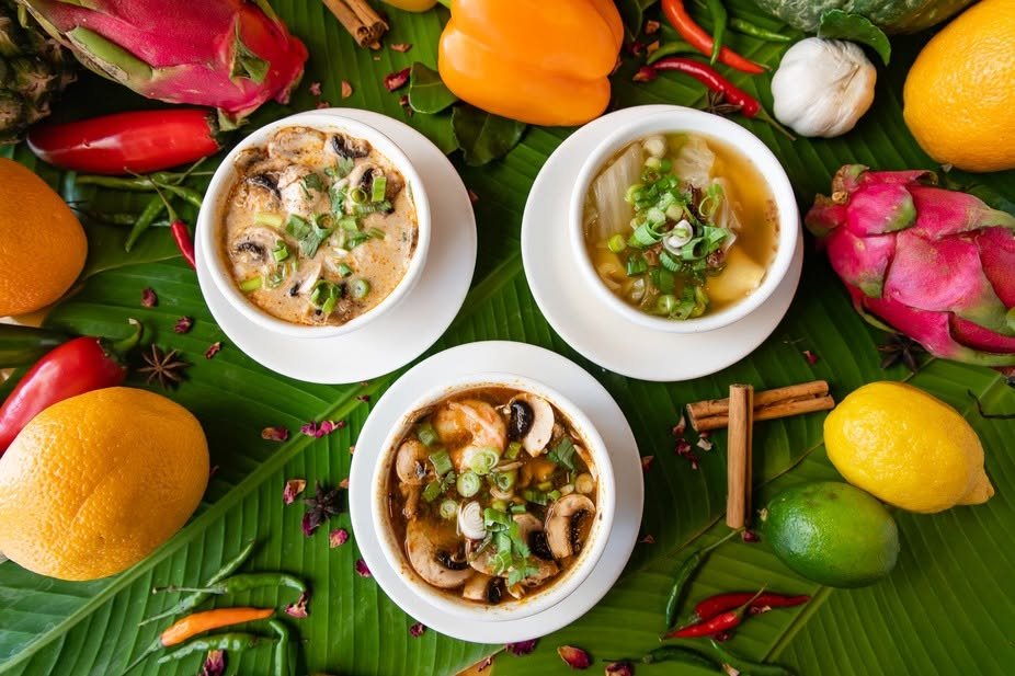 Three bowls of soup garnished with green onions on a bed of banana leaves, surrounded by various fruits and chili peppers.