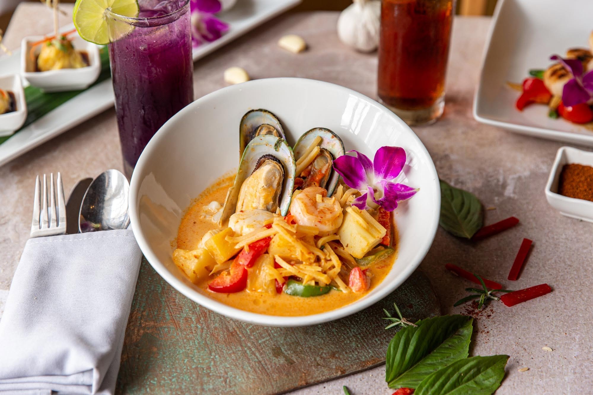 A bowl of Thai yellow curry with shrimp, vegetables, and noodles, garnished with a purple orchid flower. Surrounding the bowl are a tall glass of purple drink with a lime wedge, a glass of iced tea, and various small dishes, all on a wooden table wit