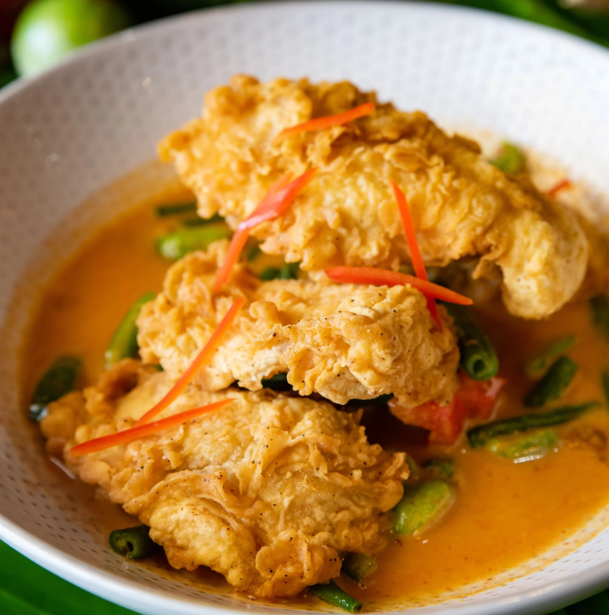 Bowl of Thai curry with a grilled fish fillet, sliced avocado, cashews, and vegetables in a orange-colored sauce.