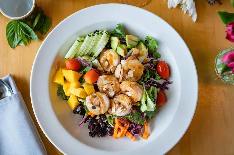 A white plate with shrimp salad containing mixed greens, cherry tomatoes, mango chunks, cucumber slices, shredded carrots, purple cabbage, avocado pieces, and blueberries.