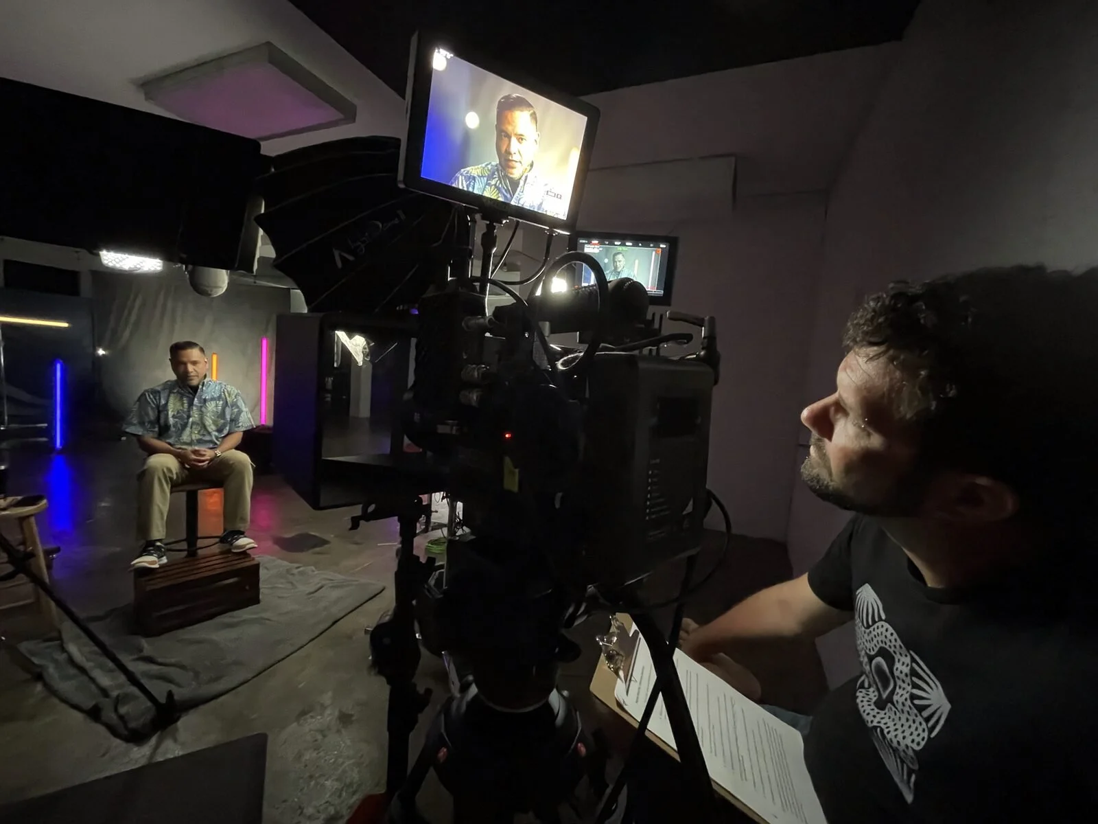 Man in a black T-shirt with a white design on it, operating camera equipment and watching a monitor, filming a man in a patterned shirt seated on a chair on a stage with colored lights.