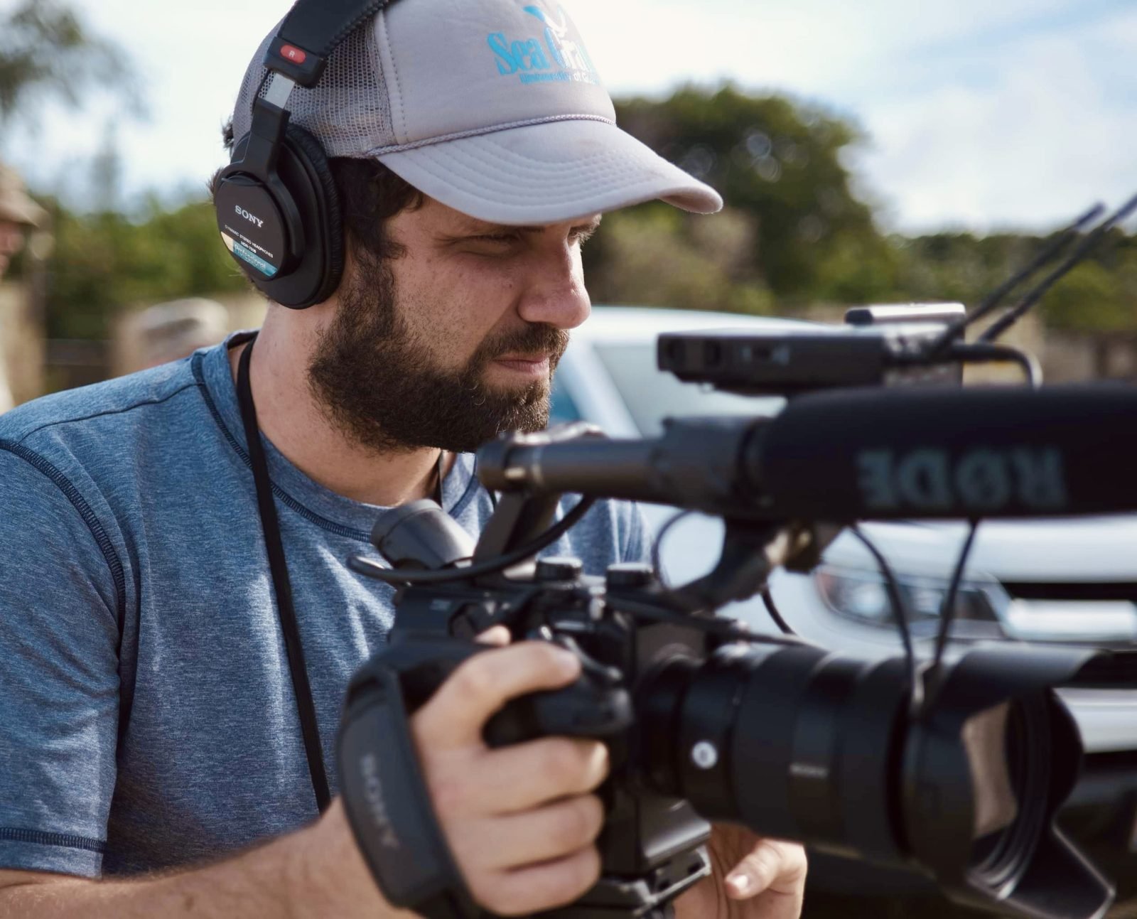 A man with a beard and a cap wearing headphones is operating a professional video camera outdoors.