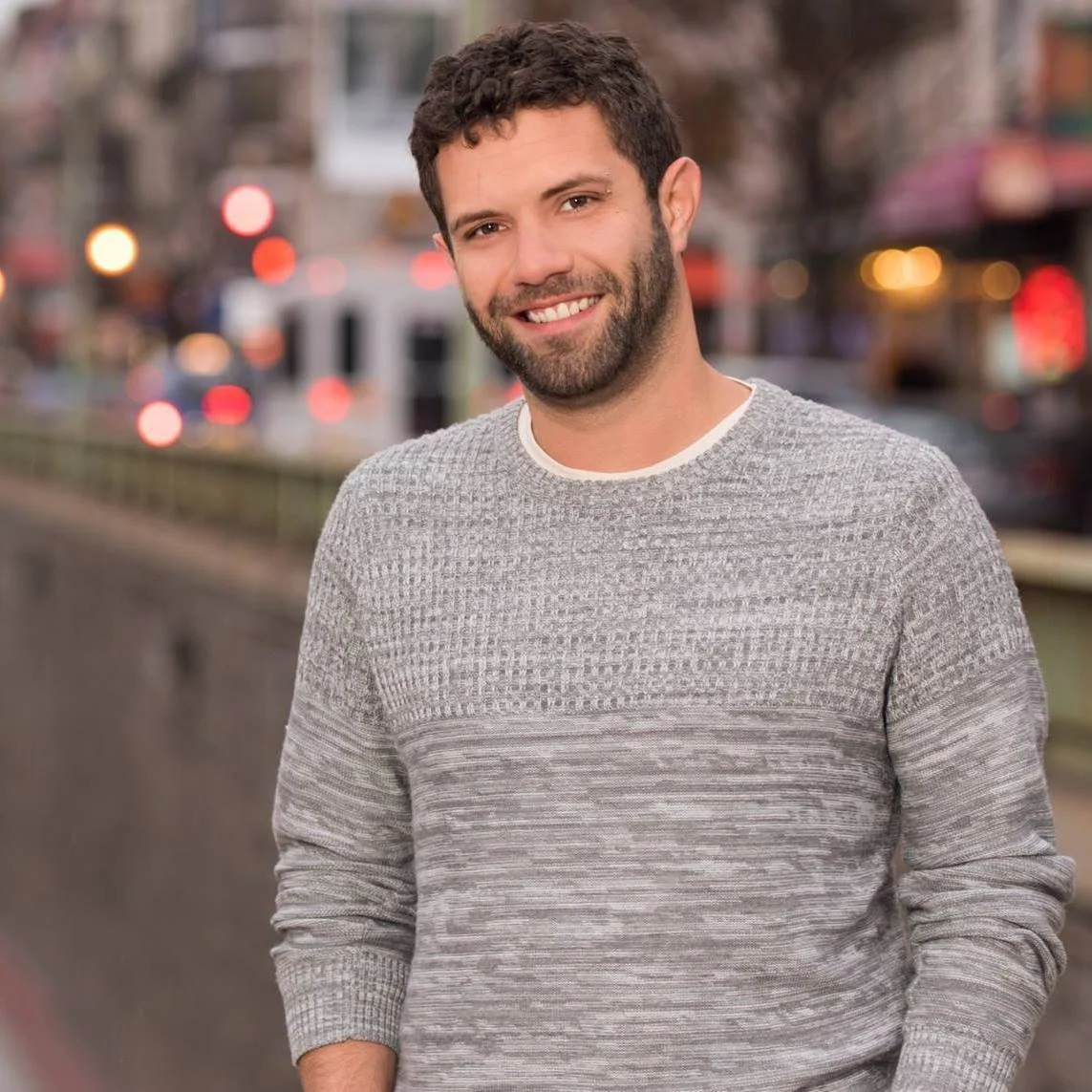 A smiling man with dark curly hair and a beard, wearing a light gray sweater, standing outdoors in an urban area with blurred city lights in the background.