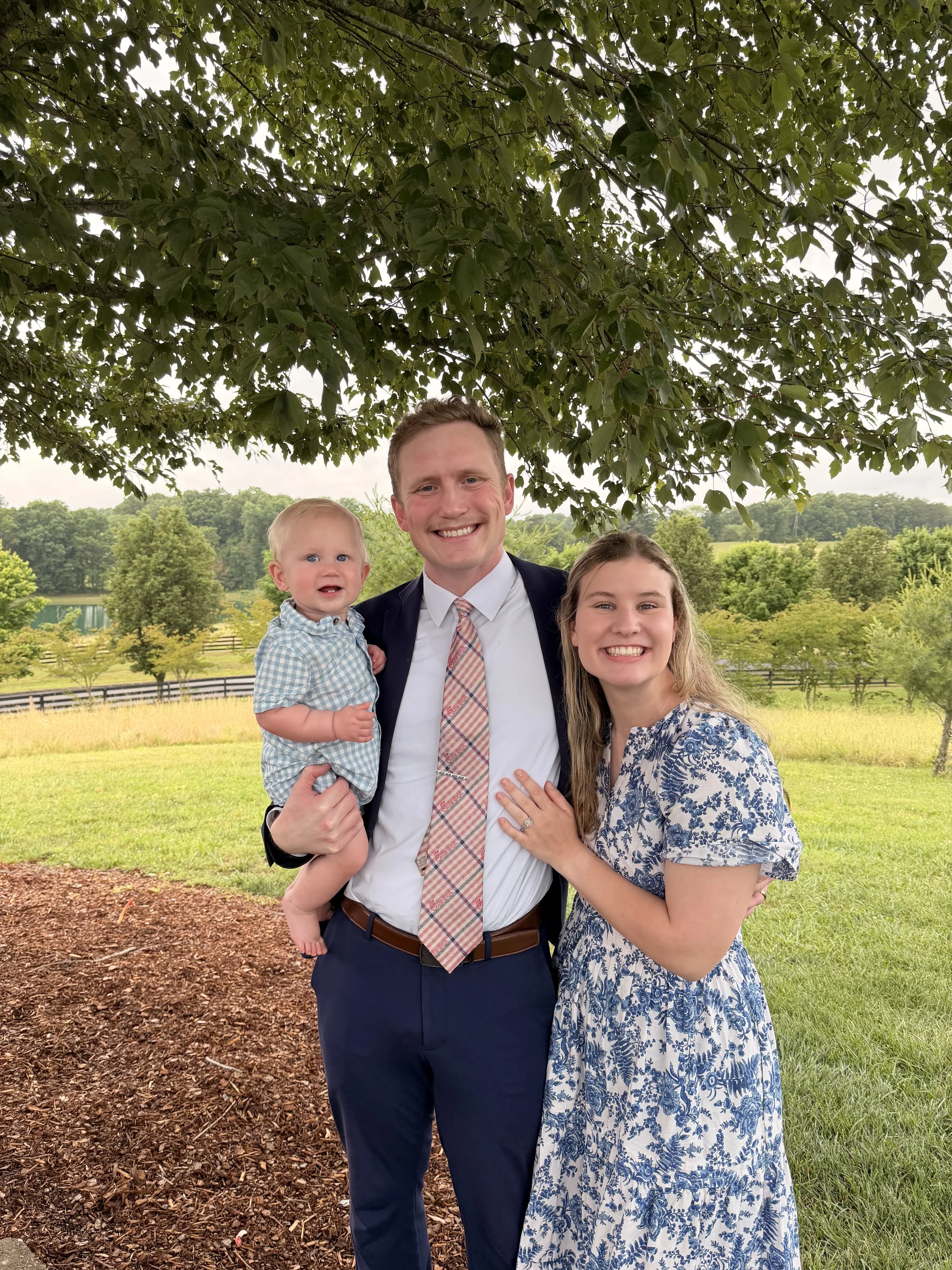A family of three poses outdoors under a large tree with green leaves, smiling at the camera. The man is holding a young boy; the woman is standing beside them. The background features lush greenery and a fence.