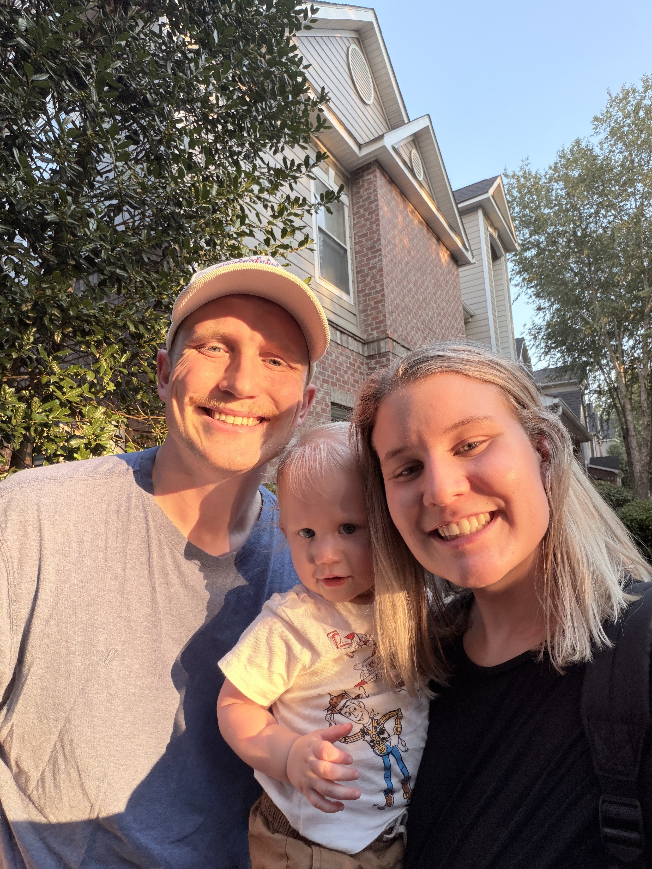 A smiling man wearing a white cap stands next to a woman with blonde hair holding a young girl with blonde hair outside a house with brick and siding walls and trees in the background.