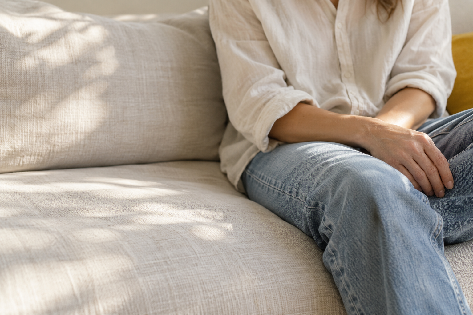Person sitting on a beige sofa, wearing a white shirt and blue jeans, with hands resting on their lap, in a sunlit room.
