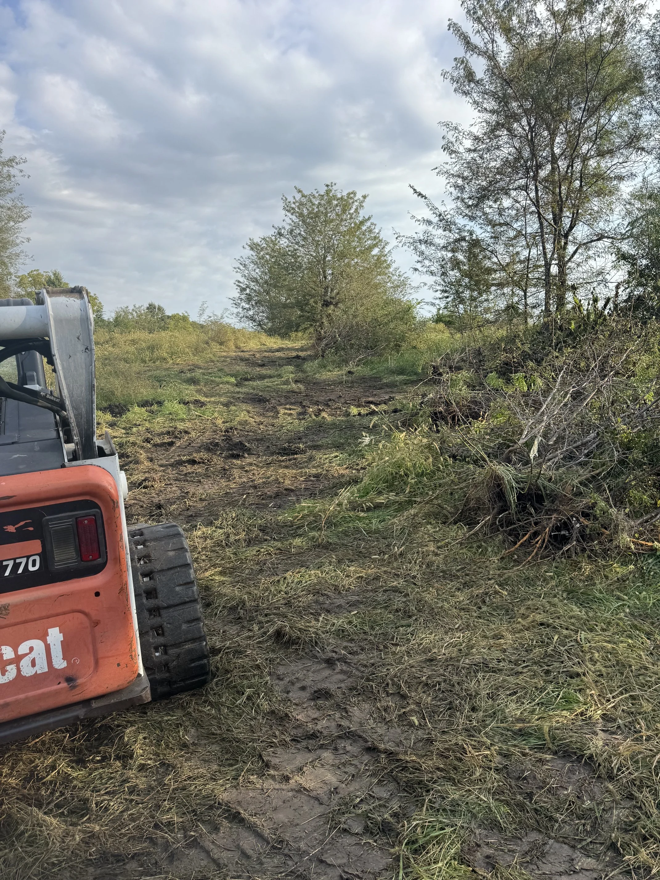 A muddy dirt road with patches of grass, surrounded by trees and bushes, with a cloudy sky overhead, and part of construction equipment visible on the left side.