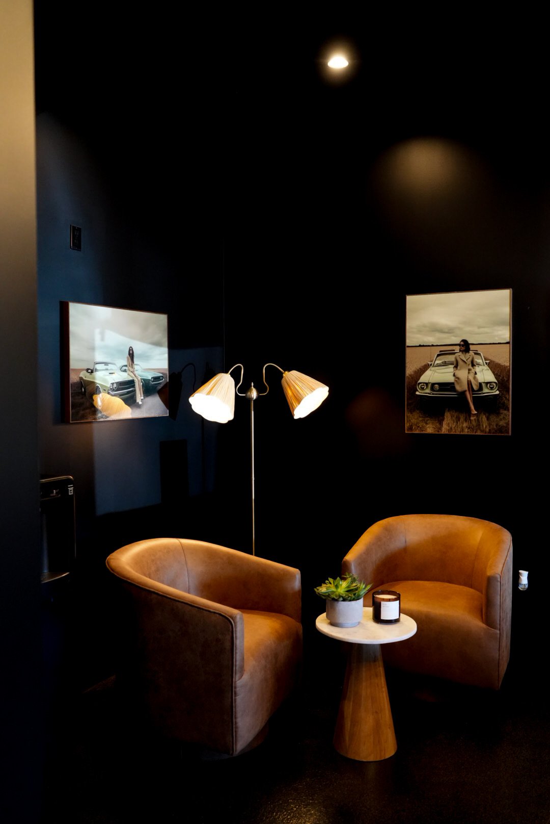Cozy seating area with two brown leather armchairs, a small round white table with a plant and candle, black walls with framed photos of women and classic cars, and a standing lamp with two shades.