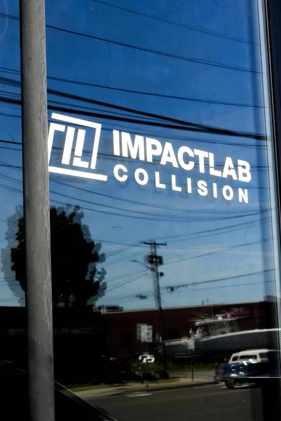 Sign for Impact Lab Collision with white text on a glass window, reflecting power lines, cars, and trees outside.