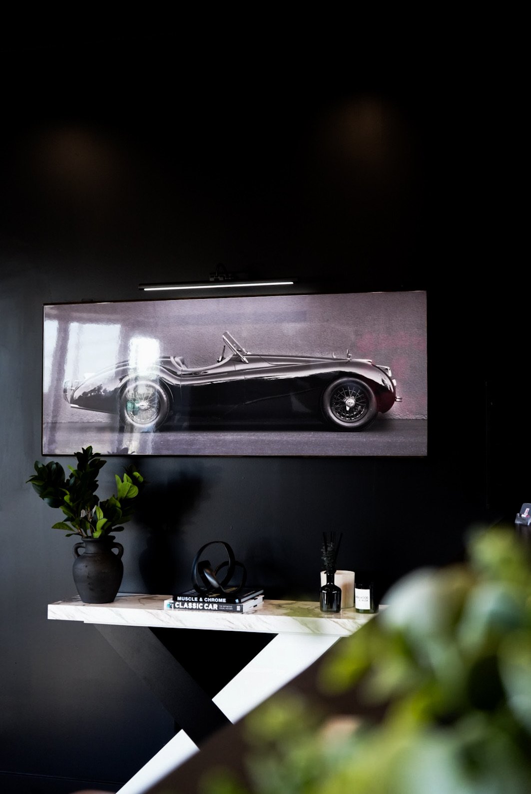 Framed black vintage sports car photograph mounted on a black wall, decorated with a potted plant, books, and decor on a white work surface, in a modern interior.