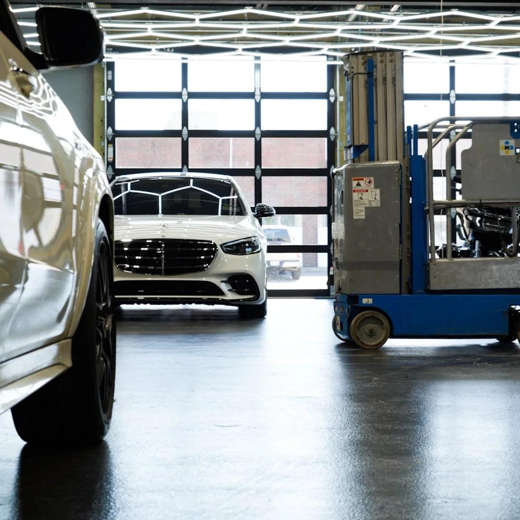 Inside a modern garage with two cars, one partially visible on the left and a white sedan in the background, along with a blue maintenance lift near the right side of the image.