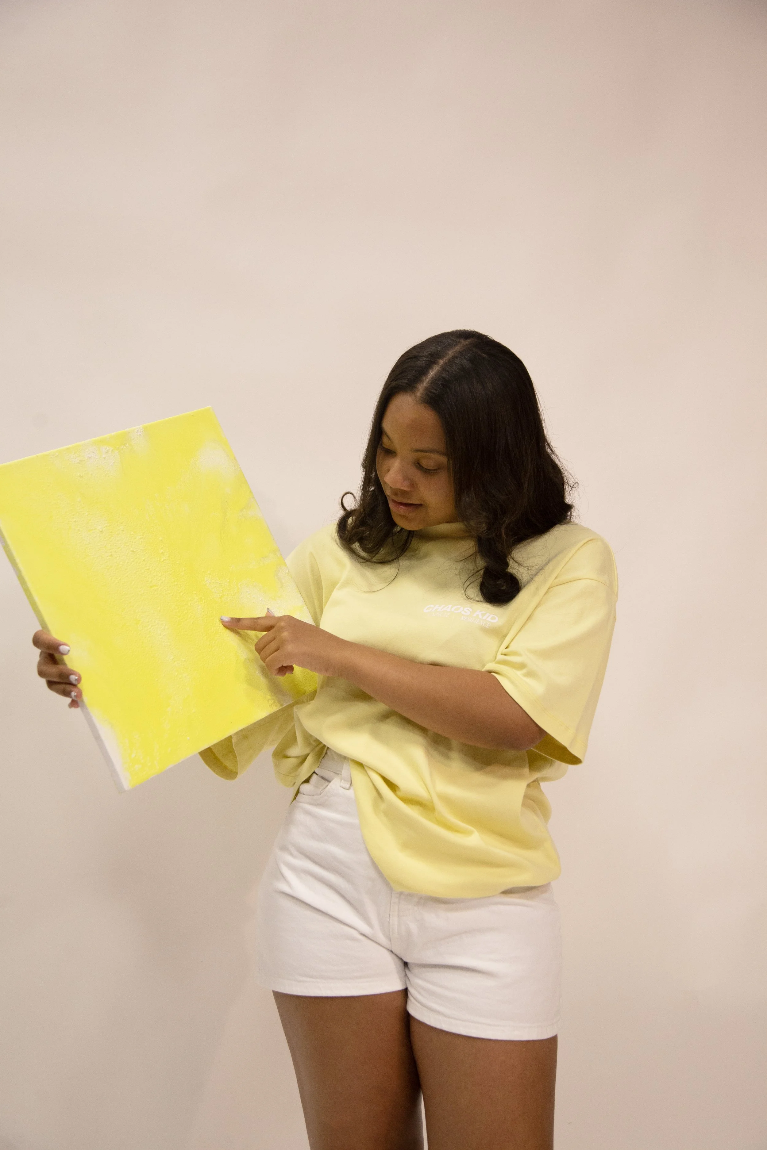 A woman in a yellow T-shirt and white shorts holding and pointing at a yellow abstract painting.