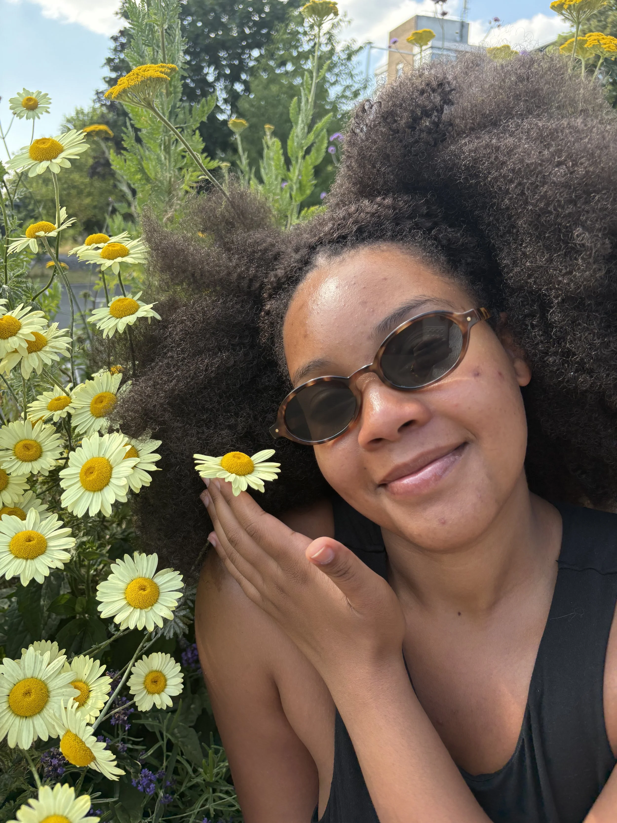 A woman with dark skin and curly hair wearing sunglasses, smiling, and touching a daisy-shaped flower with a yellow center and white petals, surrounded by yellow daisies and purple flowers in an outdoor garden.