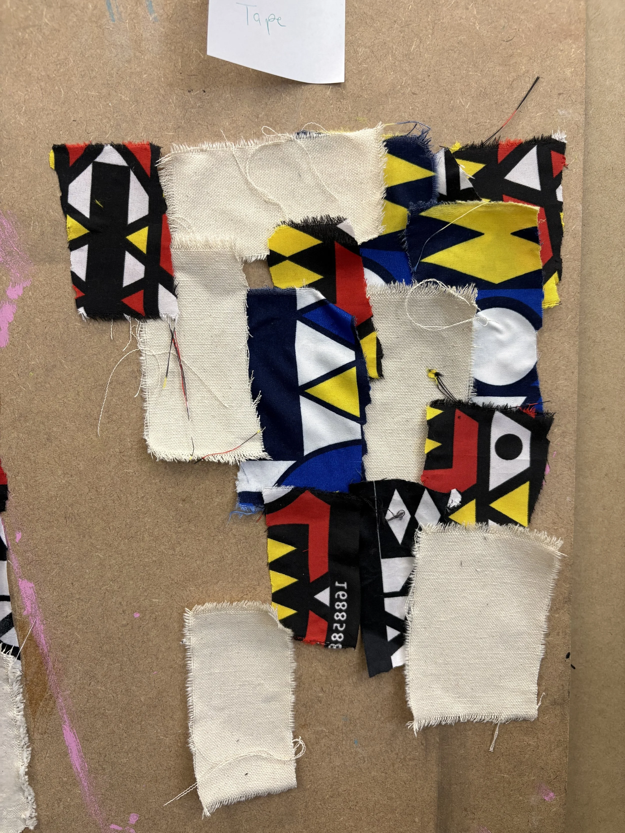 Various pieces of fabric, some with geometric patterns in black, red, yellow, and white, laid out on a brown surface with a small paper note labeled 'Tape' at the top.