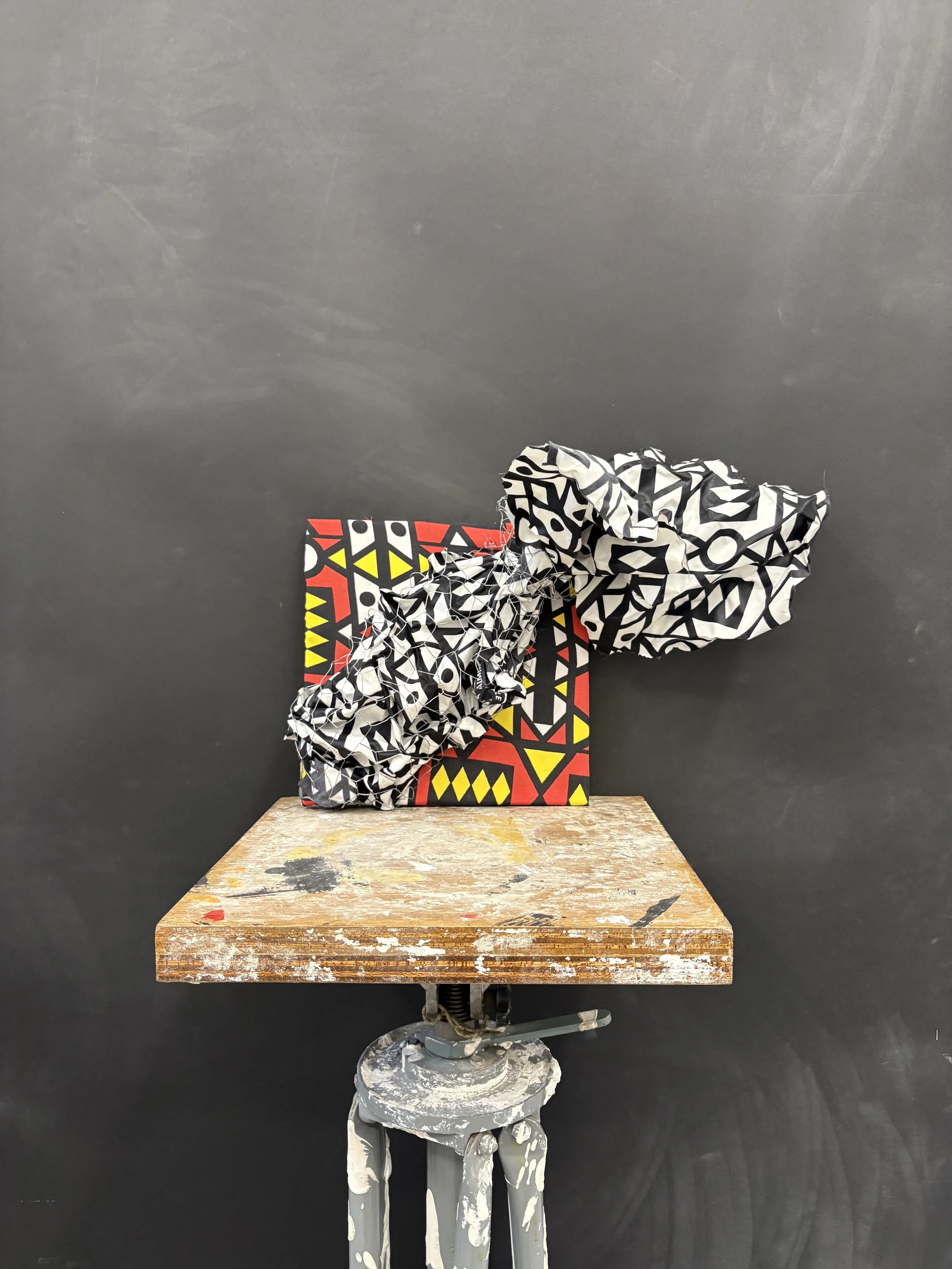 A small wooden table with paint splatters supporting an artwork consisting of a patterned box, crumpled black and white patterned paper, and intertwined wires, set against a plain black background.