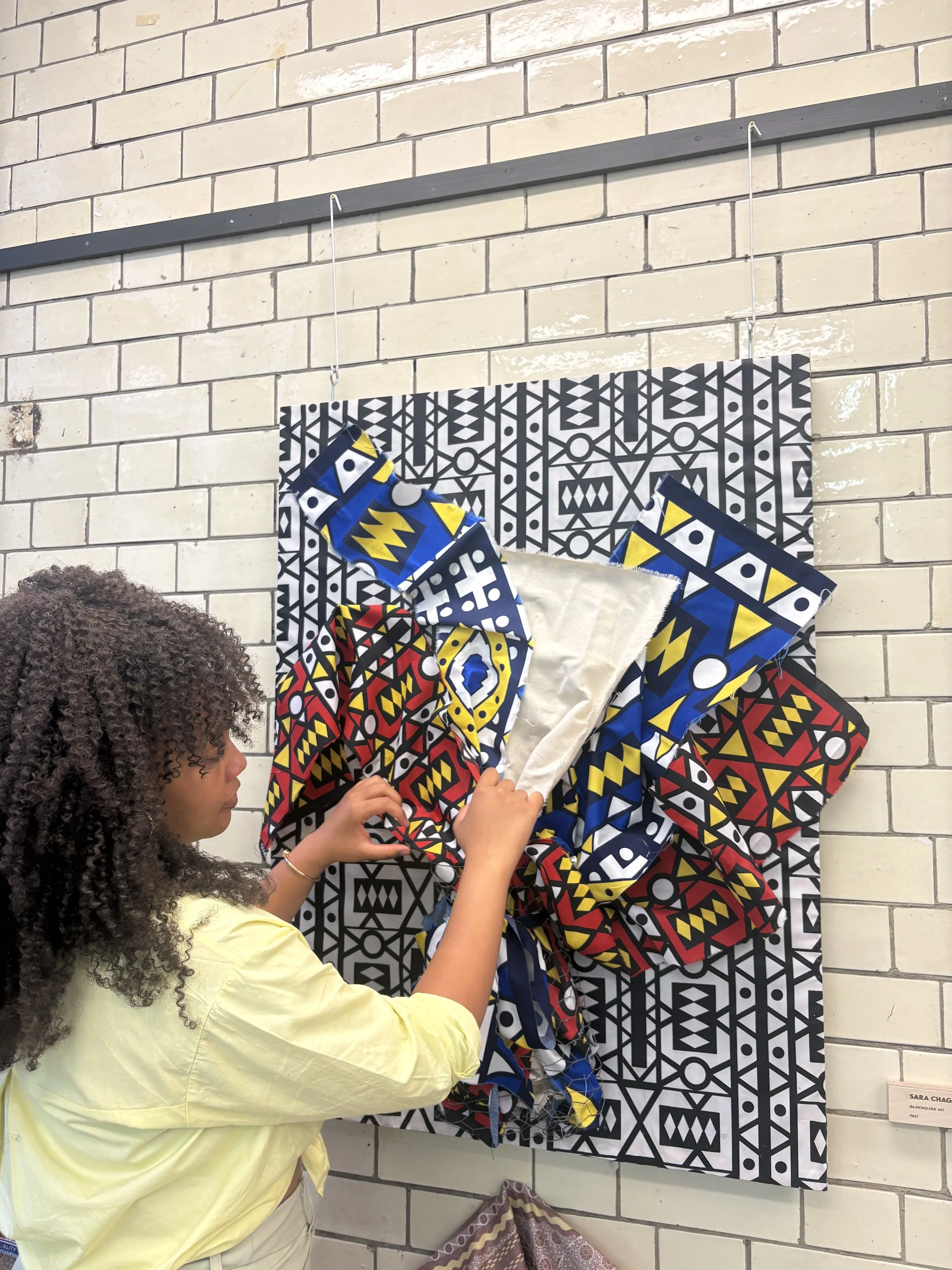 A person with curly hair wearing a yellow shirt, examining or touching a colorful, patterned textile artwork displayed on a black and white geometric patterned background. The textile has vibrant colors like red, blue, yellow, and black, and is hung in front of a white brick wall.