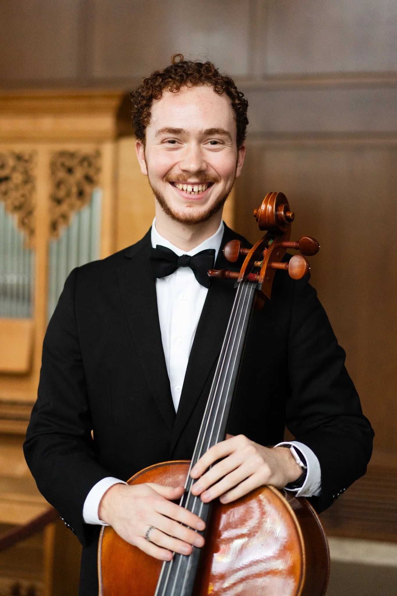 A young man with curly hair and a beard, dressed in a tuxedo, holding a cello and smiling at the camera indoors.