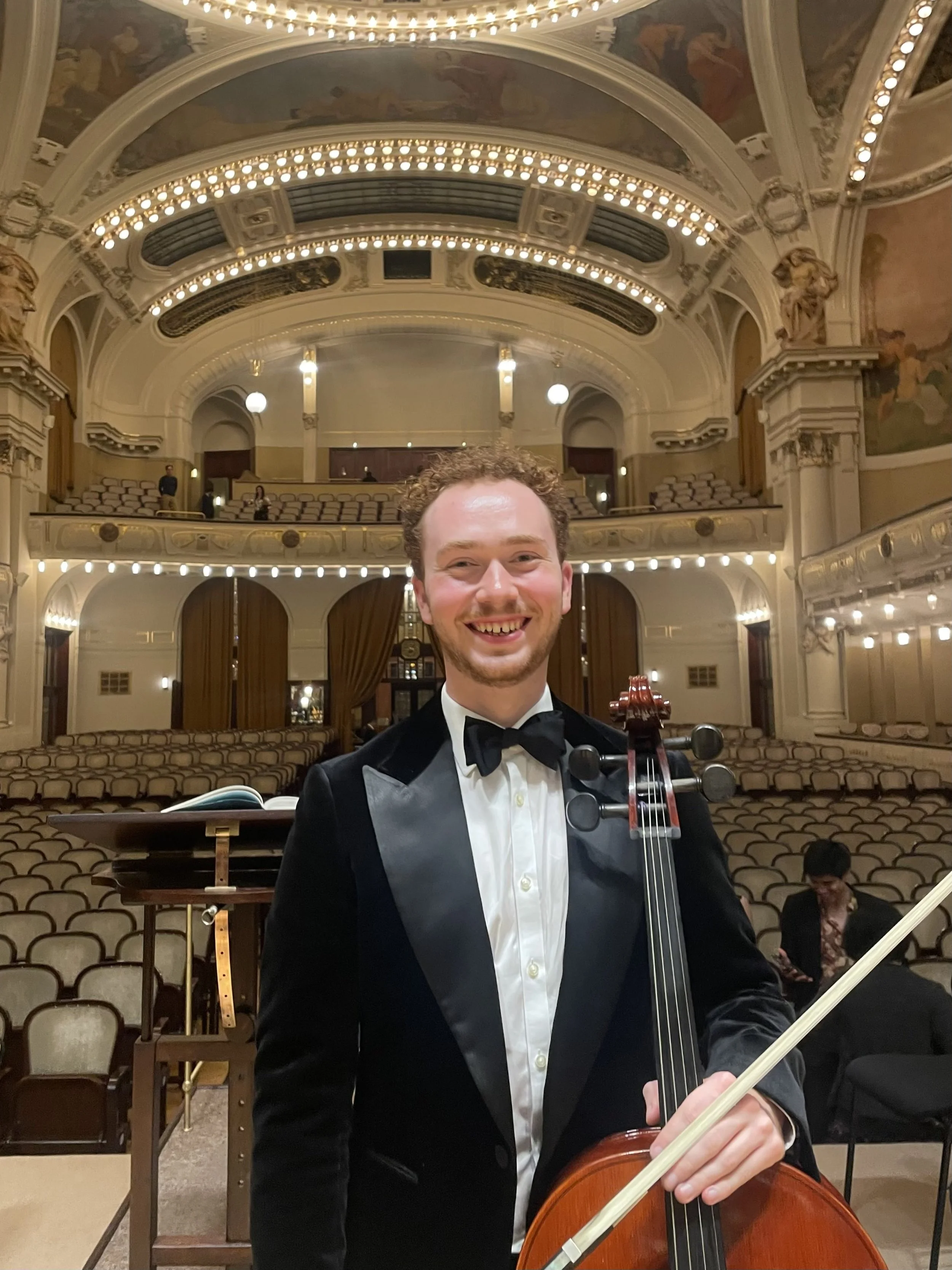 A smiling young man in formal attire holding a violin and bow on a stage in a grand concert hall with ornate architecture and empty seats in the background.