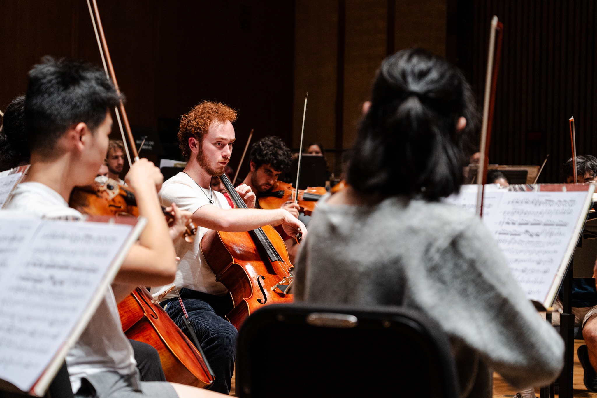 Orchestra musicians playing violins and cellos during a rehearsal in a concert hall.