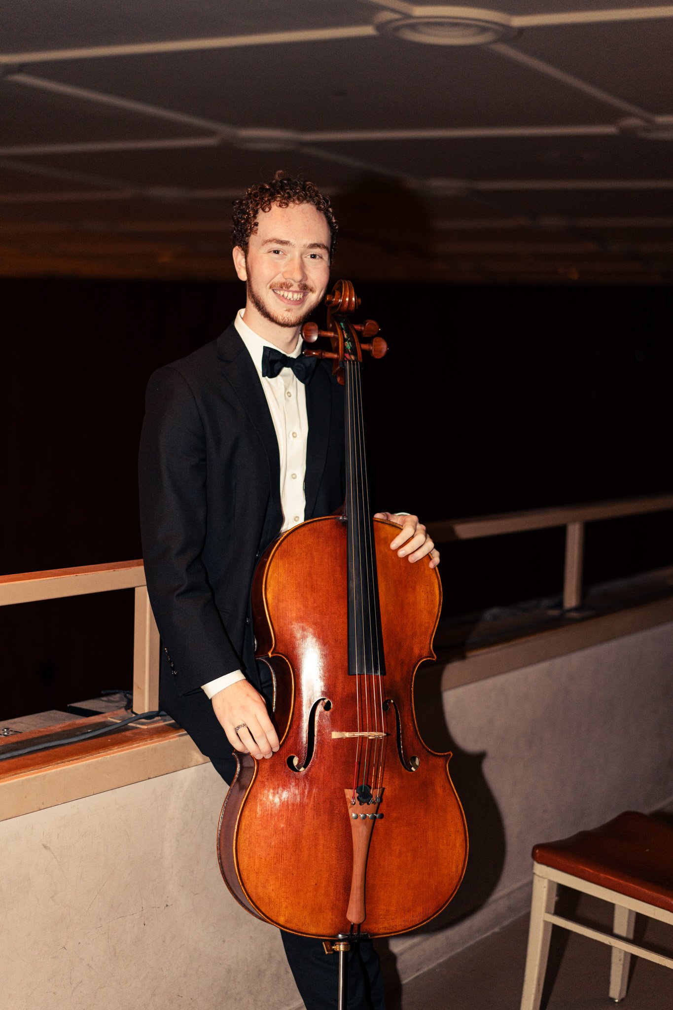 A young man with curly hair, a beard, and a black tuxedo, smiling, holding a cello in a dimly lit room.