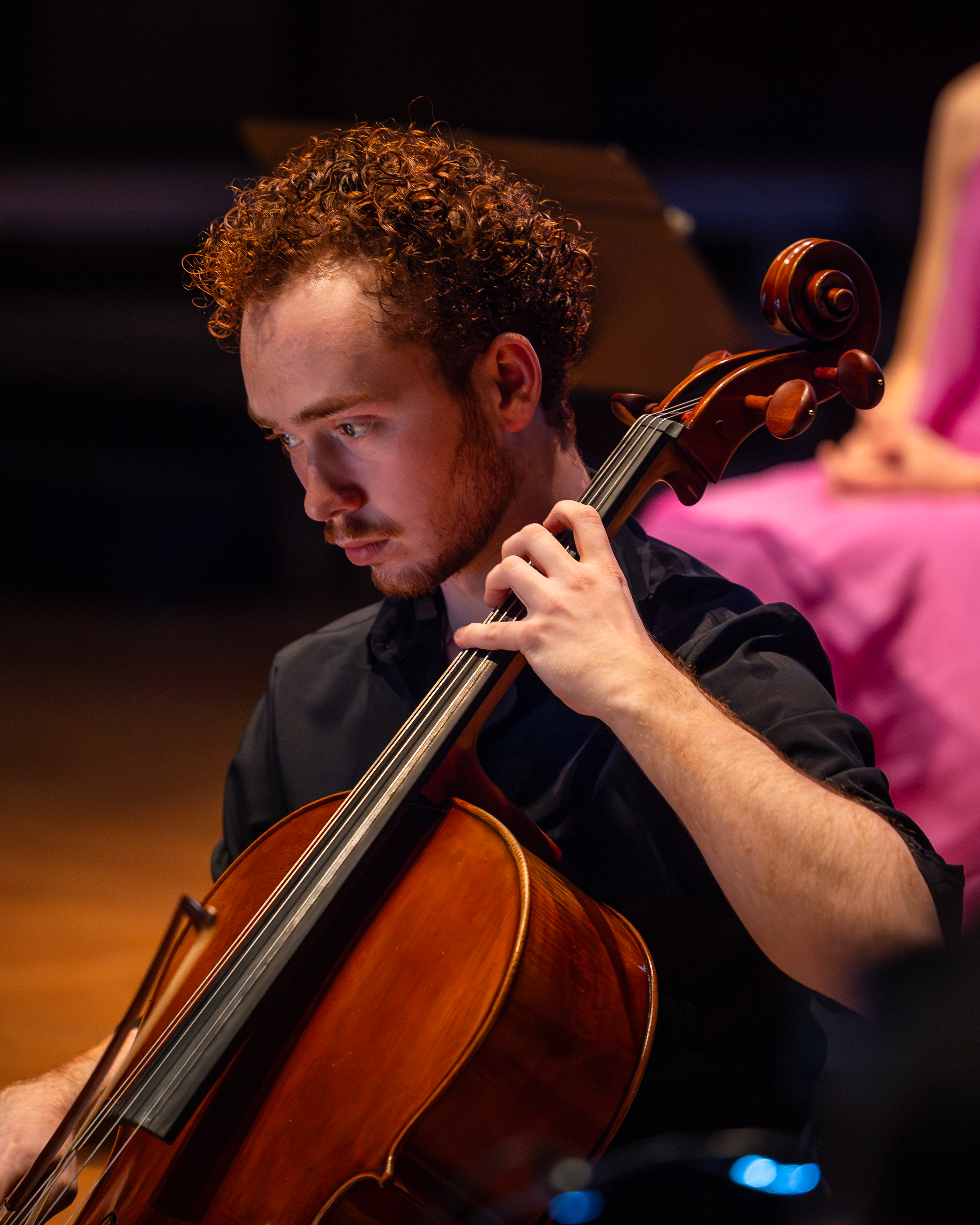 A musician playing a cello during a performance, with a focus on his face, hand, and the cello's scroll and strings, against a dark background.