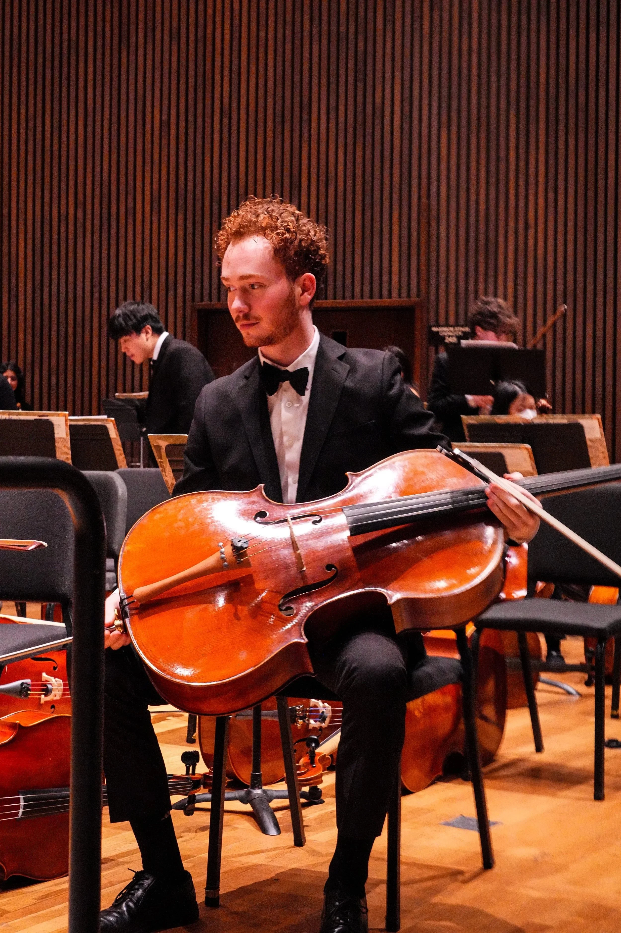 A young man with curly red hair and a beard playing a cello during an orchestra rehearsal or concert, dressed in a formal black tuxedo with a bow tie, seated in a music hall with wooden panel walls, and other musicians visible in the background.