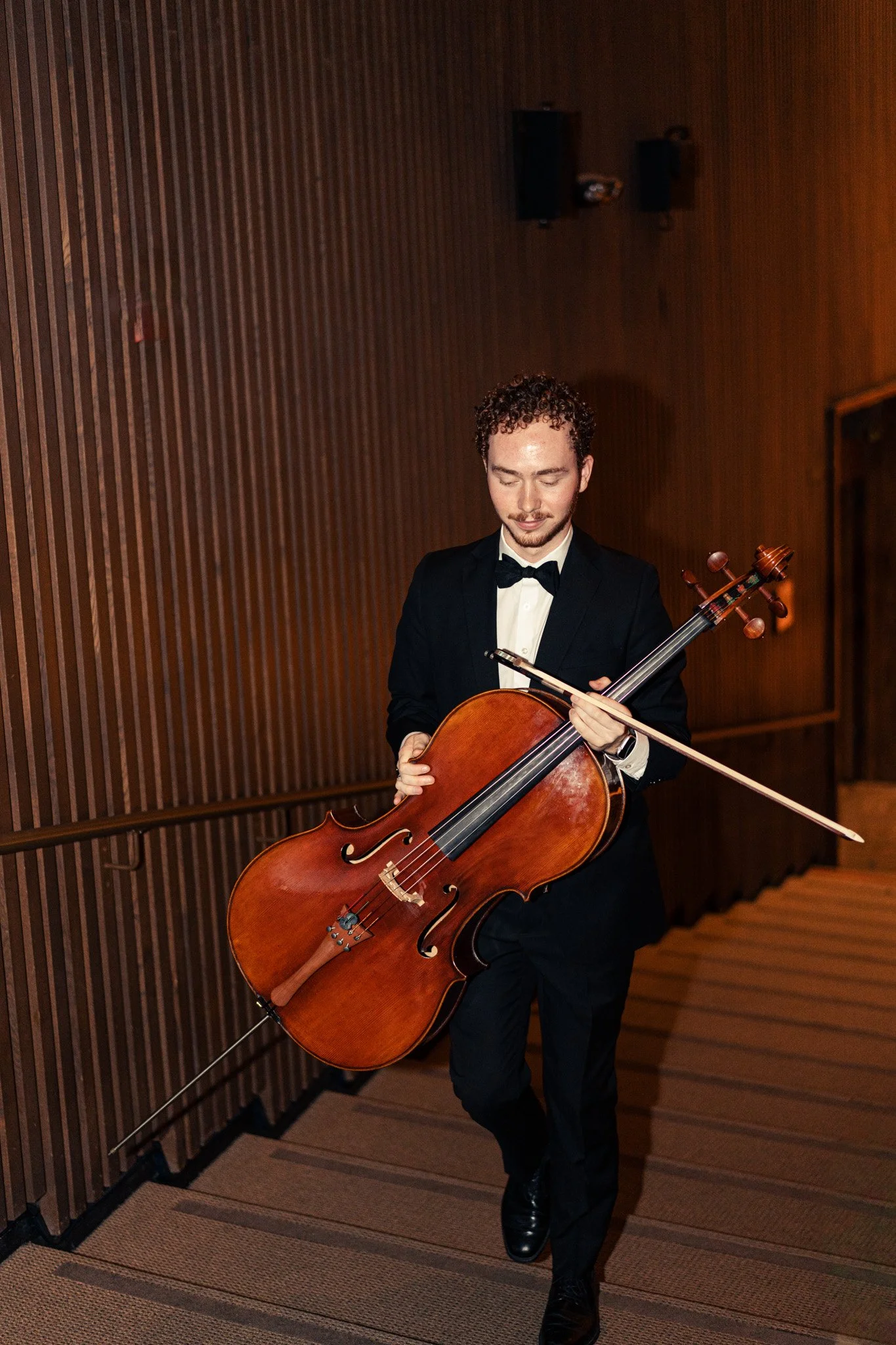 A man in a tuxedo playing a cello in a wooden-paneled room, holding a violin bow, with a focused expression.