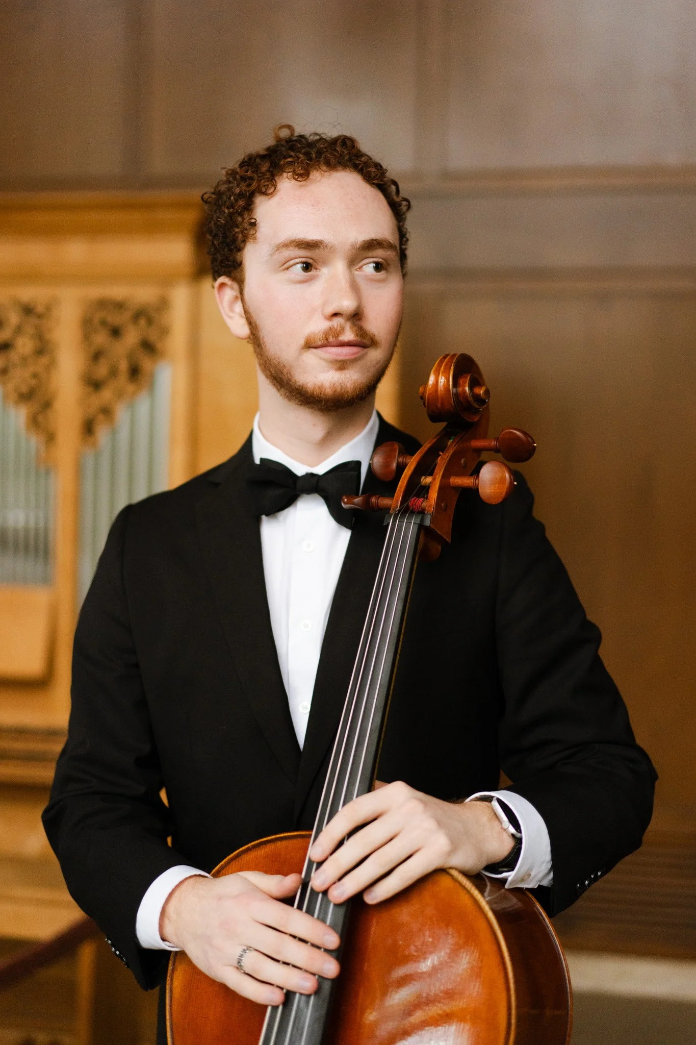 A young man in formal attire holding a cello in a room with wood-paneled walls.
