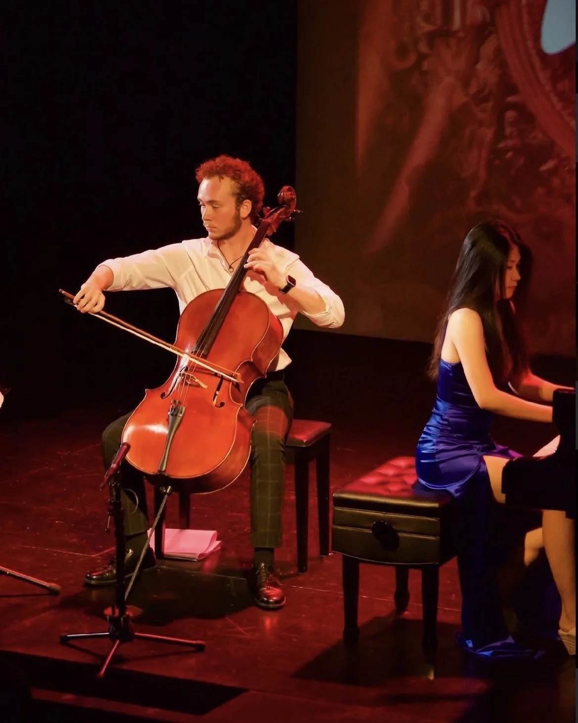 A man playing a cello and a woman playing a piano on stage during a live performance.