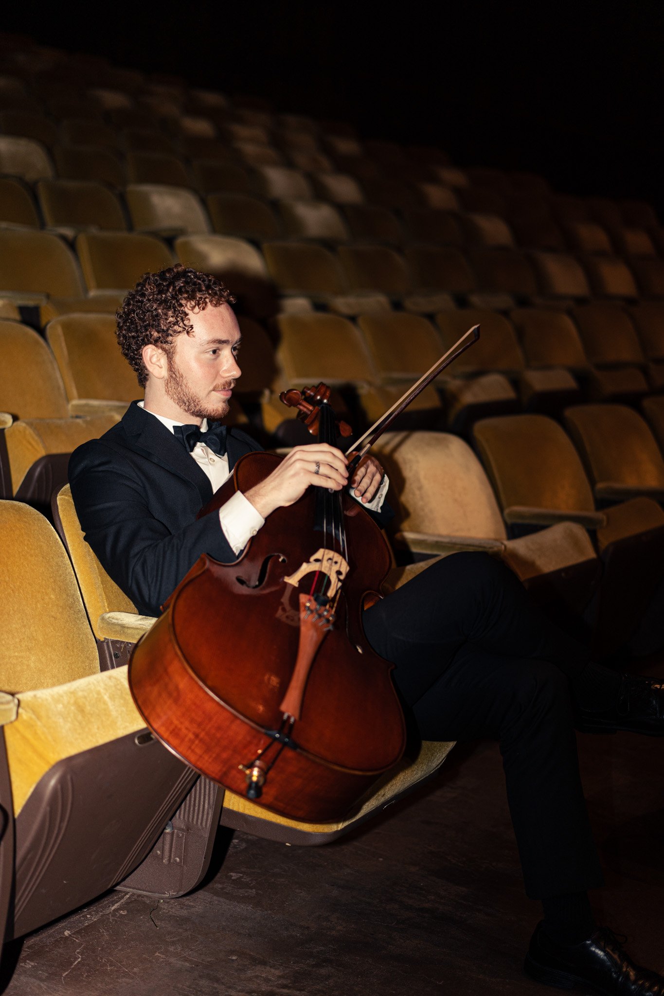 A man in a tuxedo playing a cello while sitting in an auditorium with empty yellow seats.