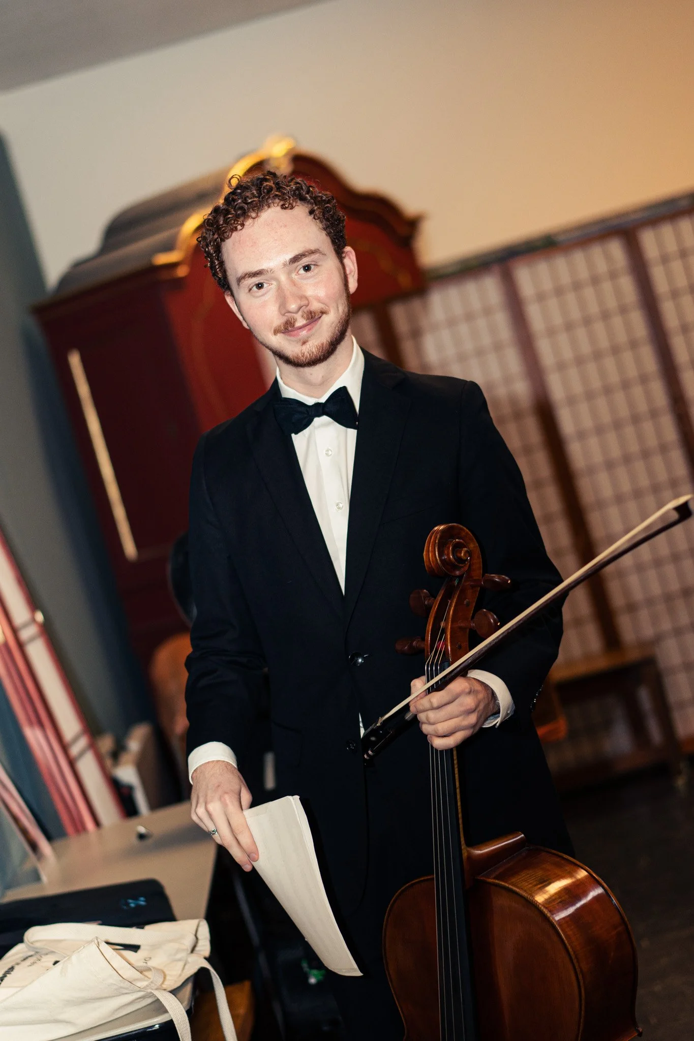 A young man with curly hair and a beard, dressed in a black tuxedo with a bow tie, stands holding a cello and a bow in a room with musical equipment and wooden decor.