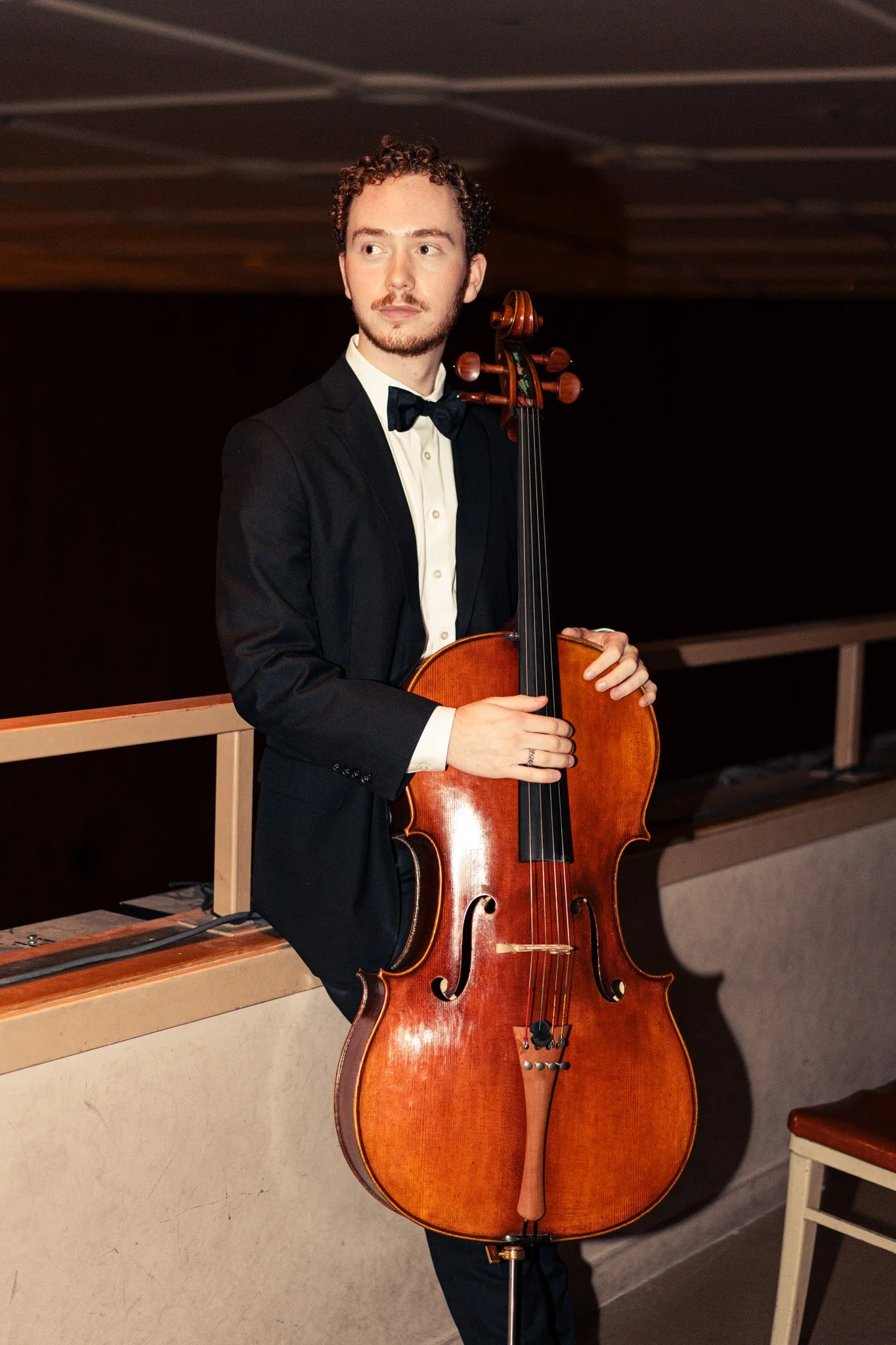A man in a tuxedo holding a cello indoors, standing by a bench and dark background.