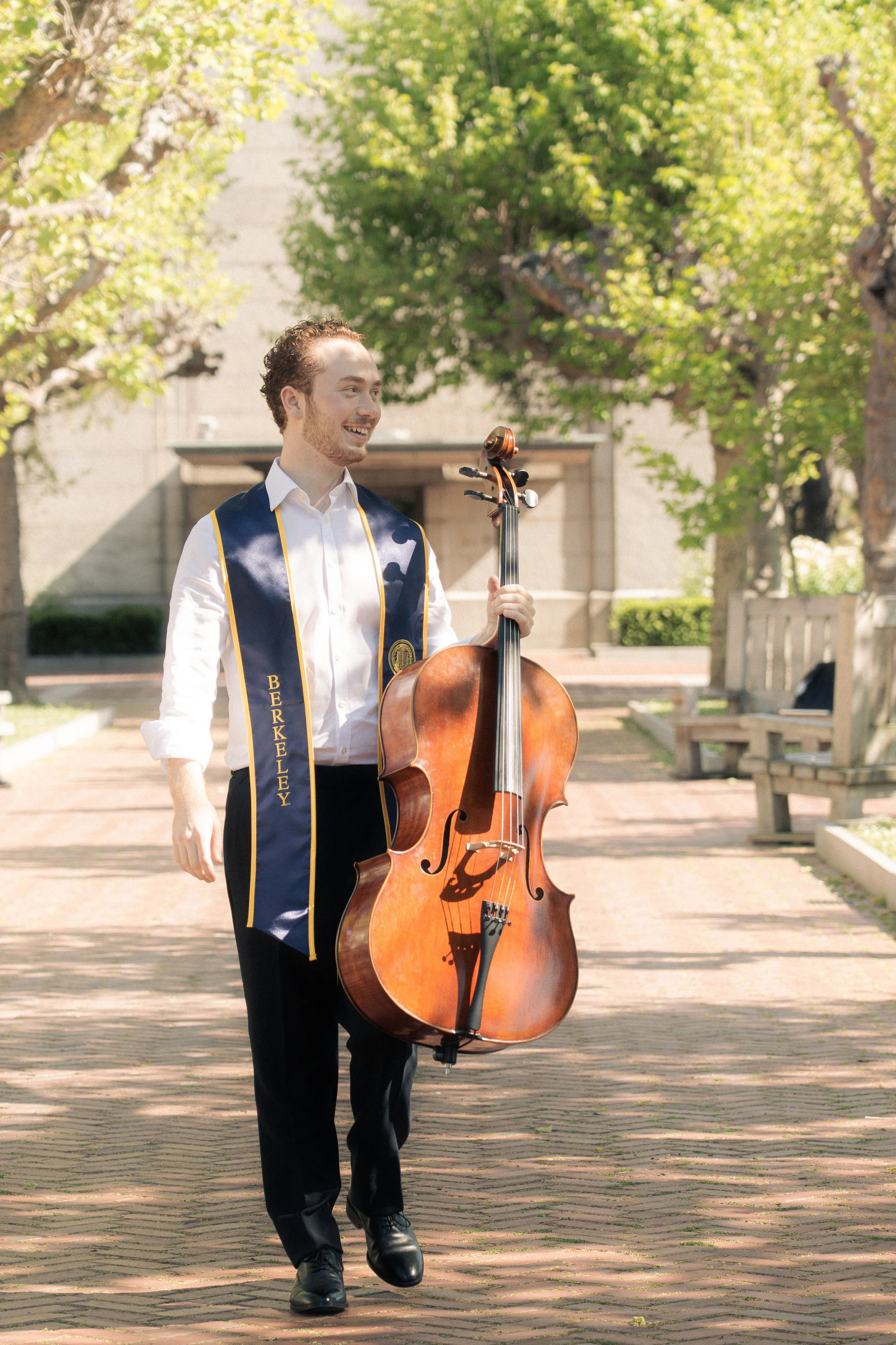 A young man dressed in formal attire, wearing a sash with 'Berkeley' on it, is smiling and holding a cello in an outdoor park setting with trees and benches.