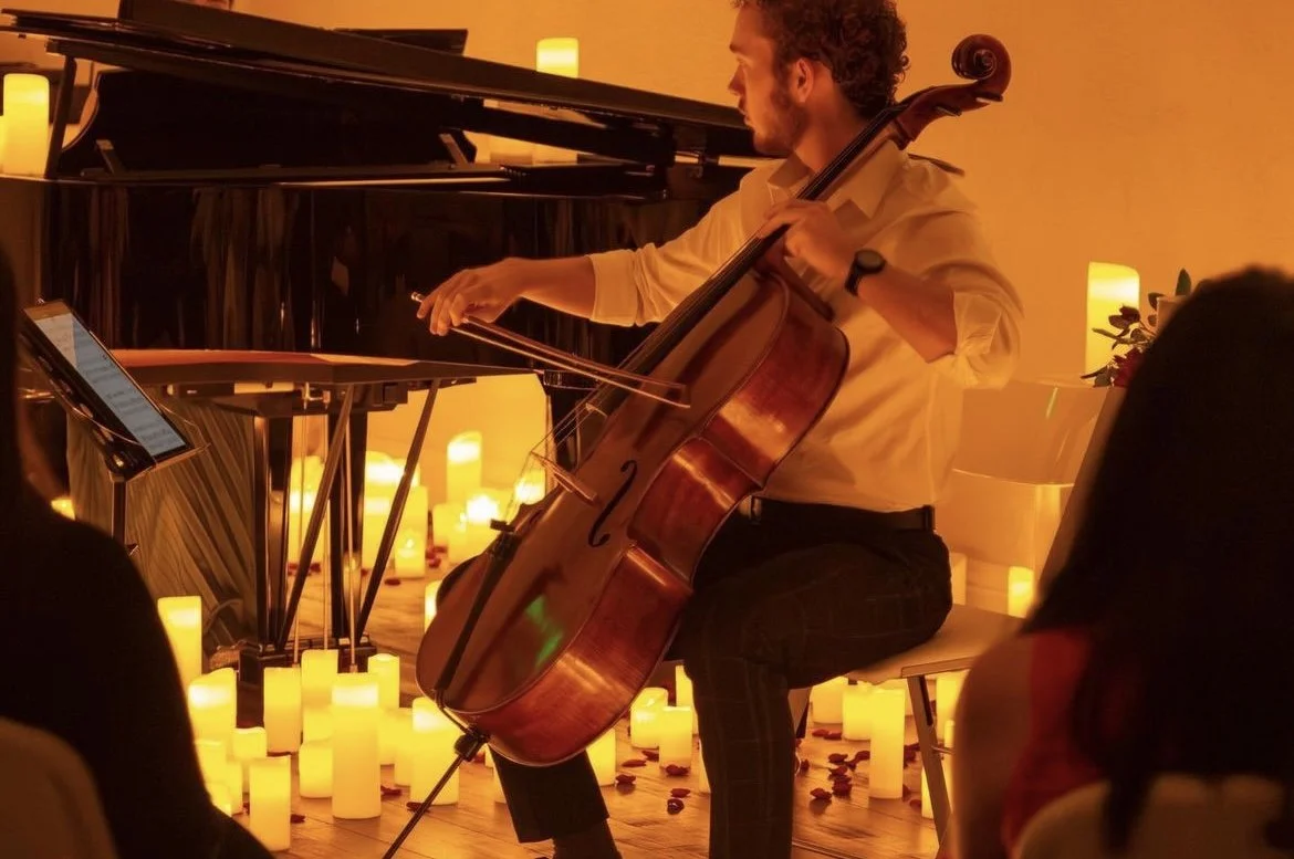 A man playing a cello during a performance surrounded by numerous lit candles, with a black grand piano in the background and candles on the floor and on a table, creating a warm, intimate atmosphere.