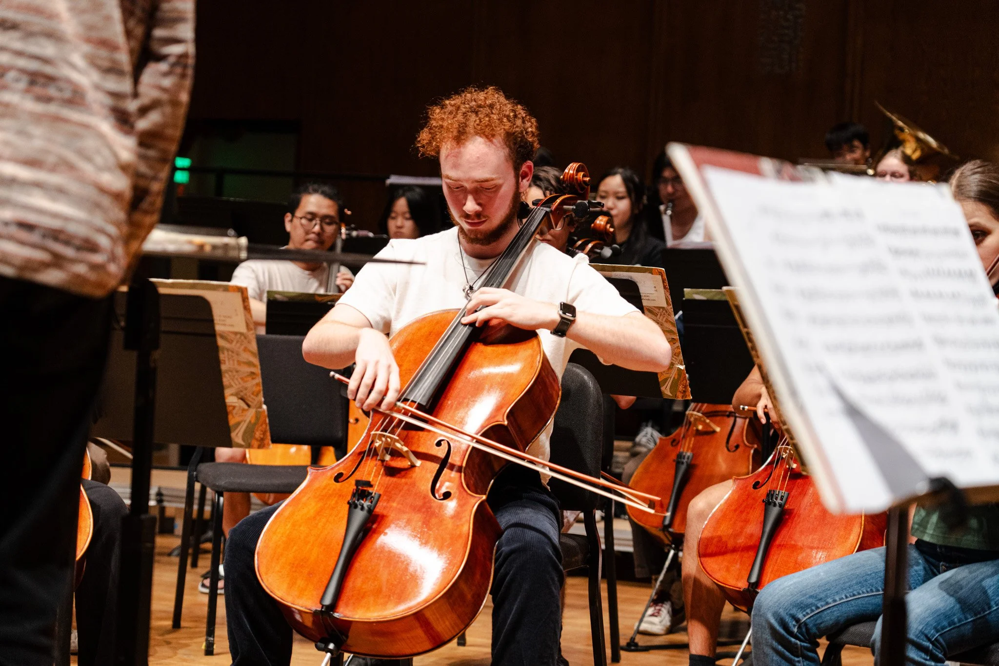 A young man with curly red hair playing a cello during a rehearsal with an orchestra in a concert hall, with other musicians and sheet music around him.