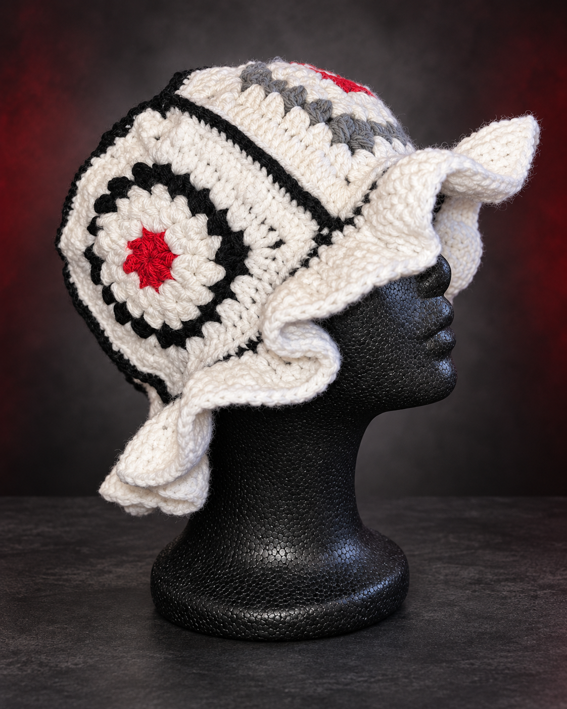 Handmade white, black, gray, and red granny square crochet bucket hat with a ruffled brim on a black mannequin head.