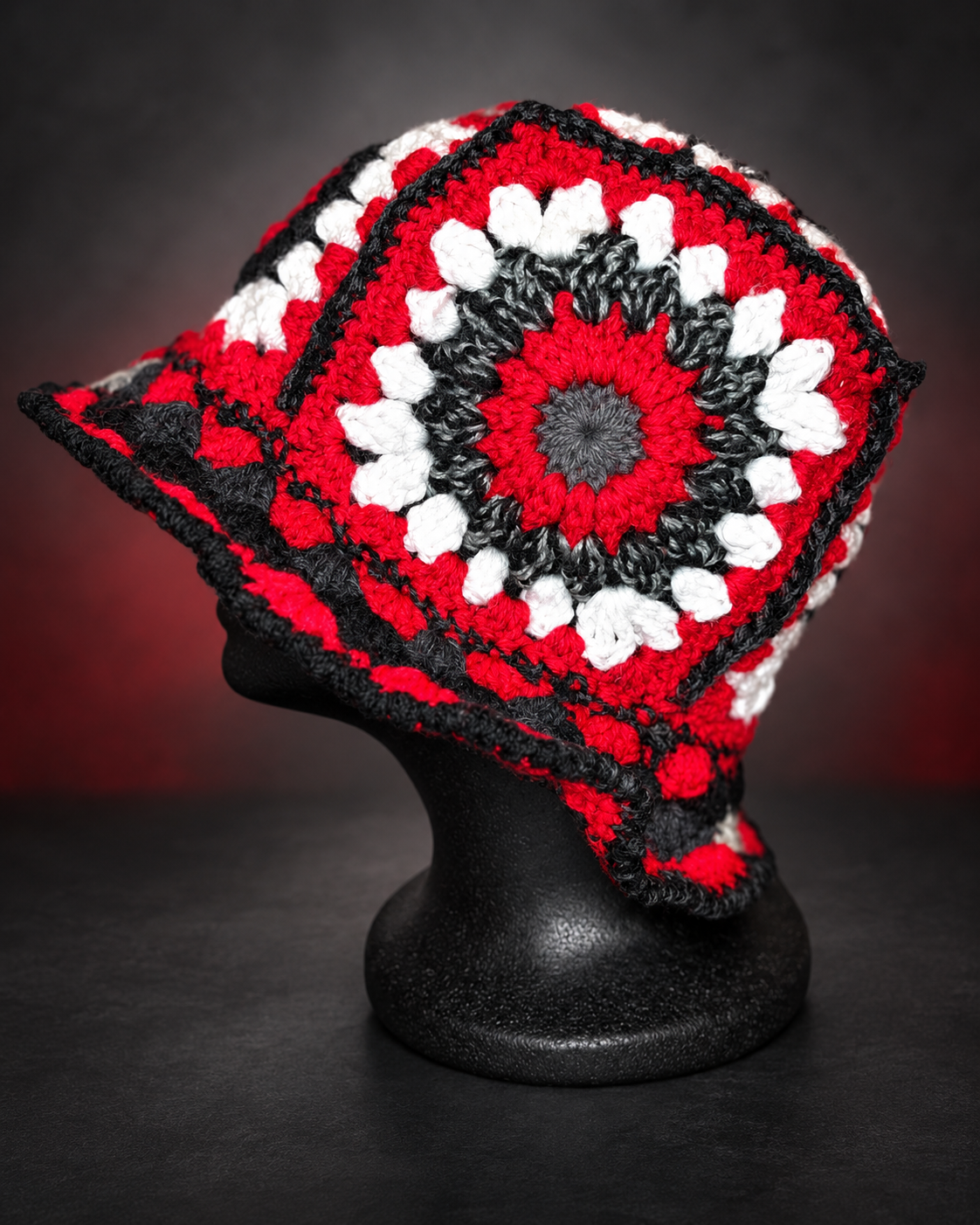 A colorful, crocheted square pattern headpiece with red, black, white, and gray colors displayed on a black mannequin head against a dark background.