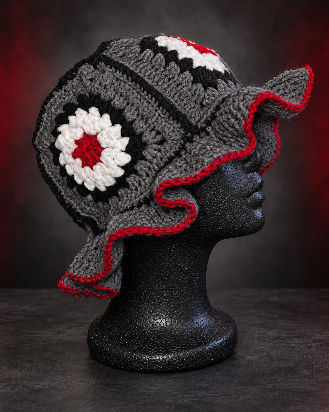 Handcrafted crocheted hat with red, black, white, and gray yarn on a black mannequin head, against dark background.