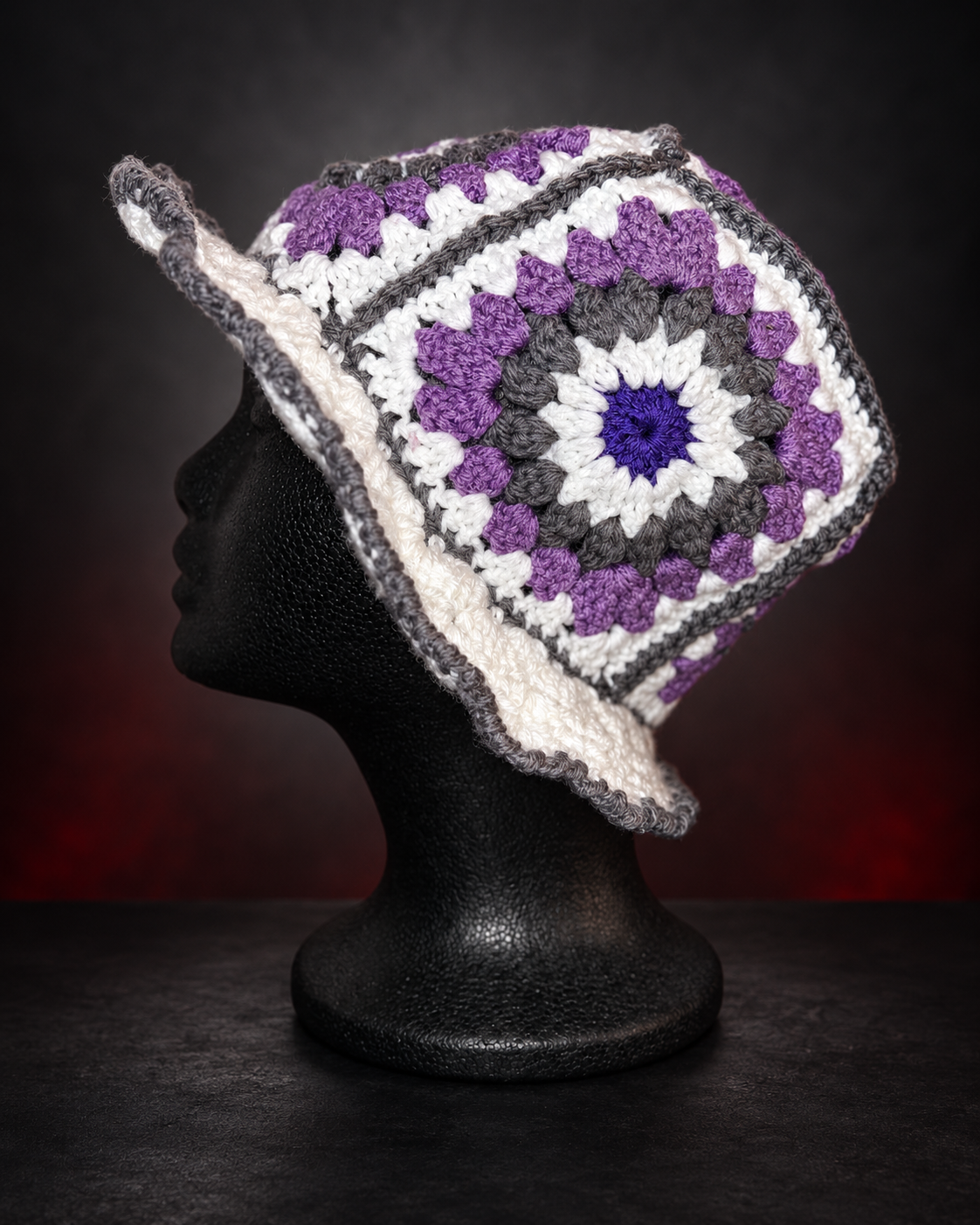 A black mannequin head wearing a colorful crochet hat with purple, white, gray, and blue yarn in a geometric pattern, against a dark background.