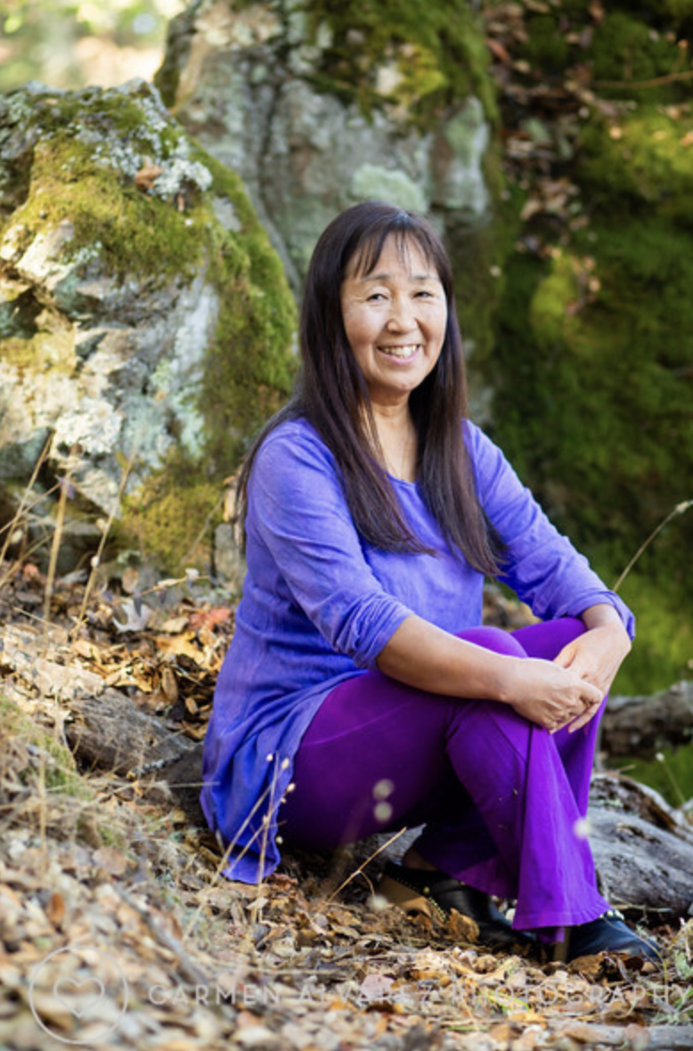 Yasue sitting in front of a rock wearing purple clothes