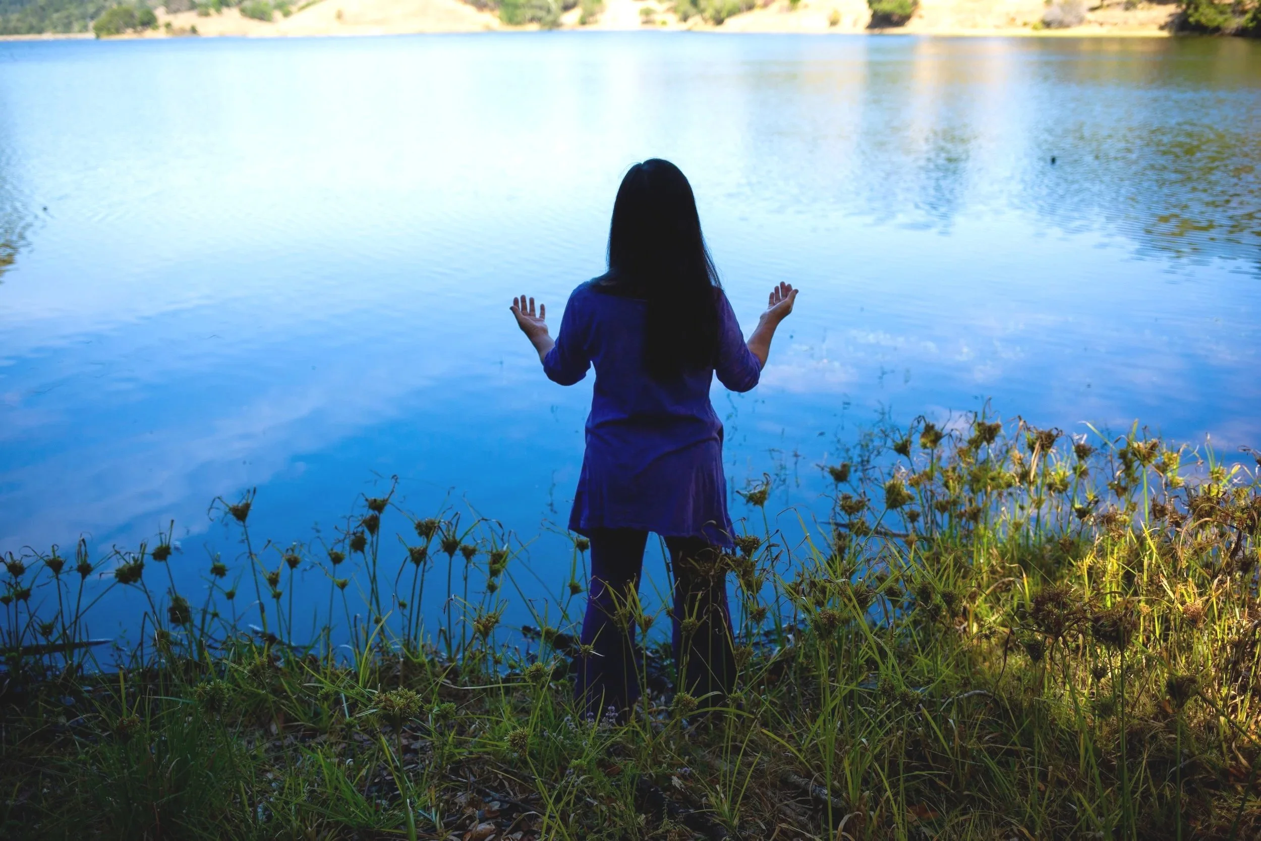 Yasue Tanaka standing at the edge of a lake with her back to the camera and her hands raised.