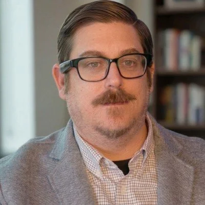 A man with brown hair, glasses, and a mustache, wearing a gray blazer and a light checkered shirt, in front of a bookshelf.