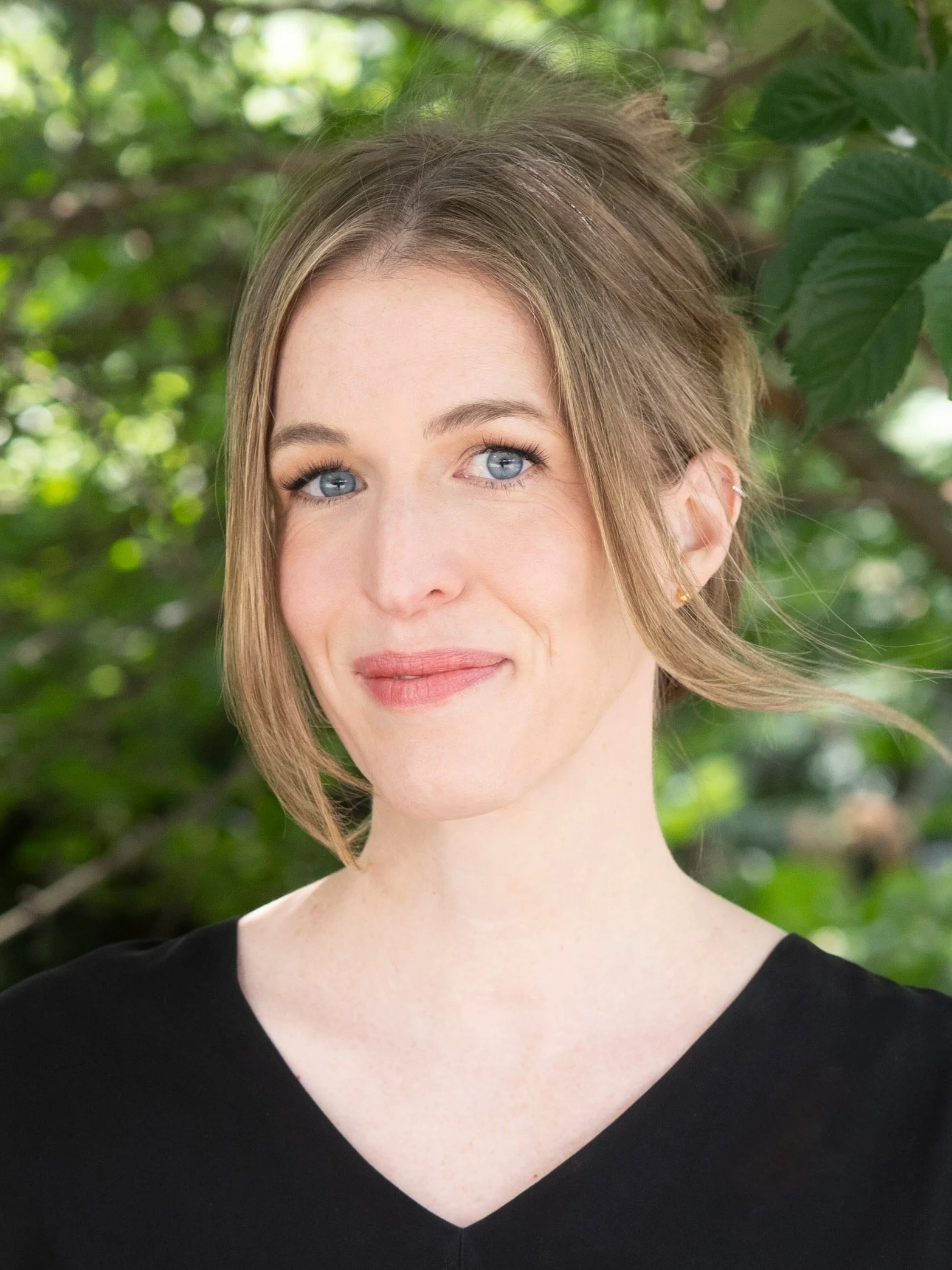 Close-up of a woman with blonde hair and blue eyes, wearing a black top, standing outdoors with green leaves in the background.