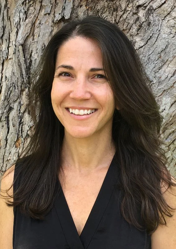 A woman with long dark hair smiling in front of a tree trunk.