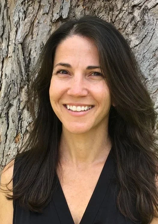 A woman with long dark hair smiling in front of a large tree trunk.