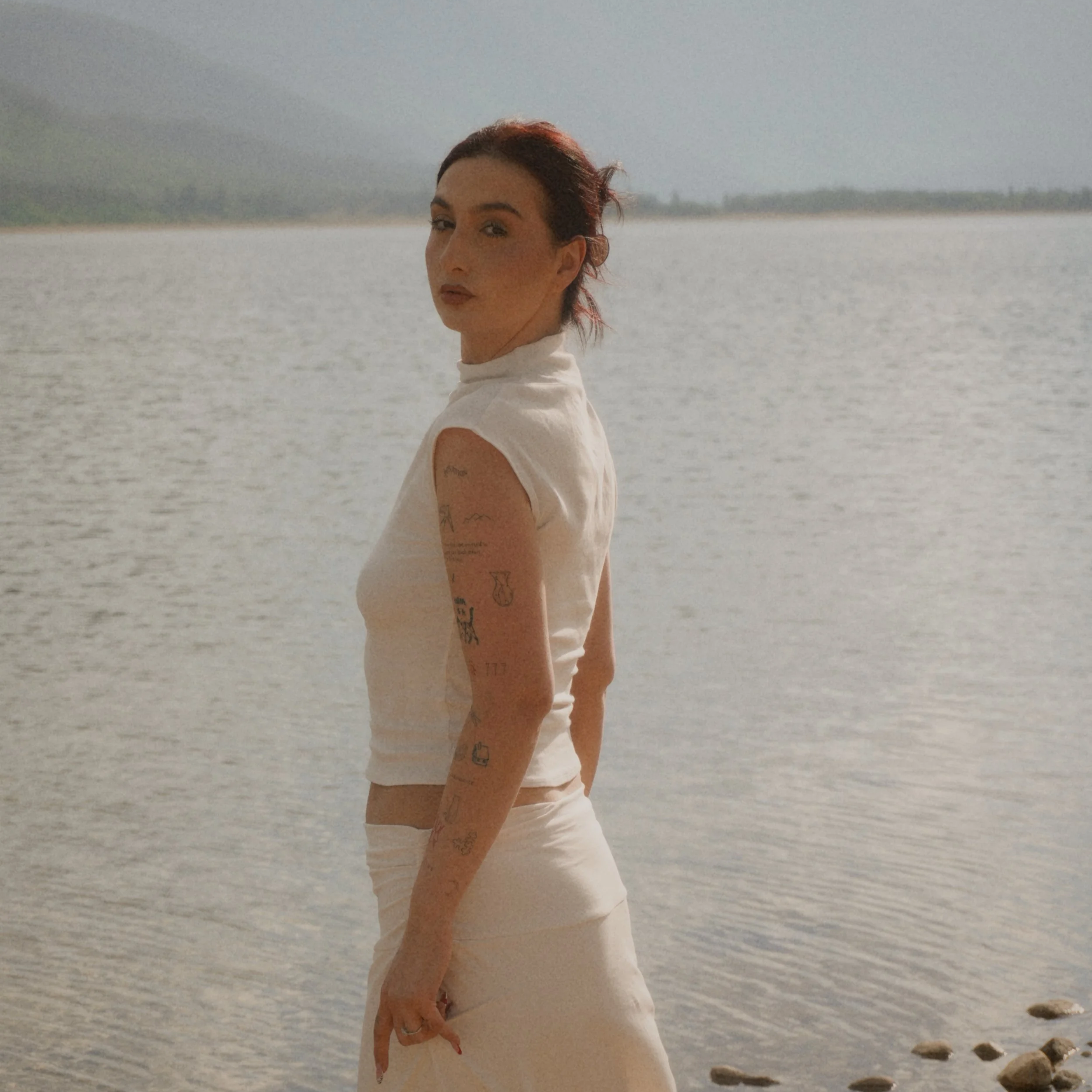 Woman with red hair and tattoos standing near water on a cloudy day