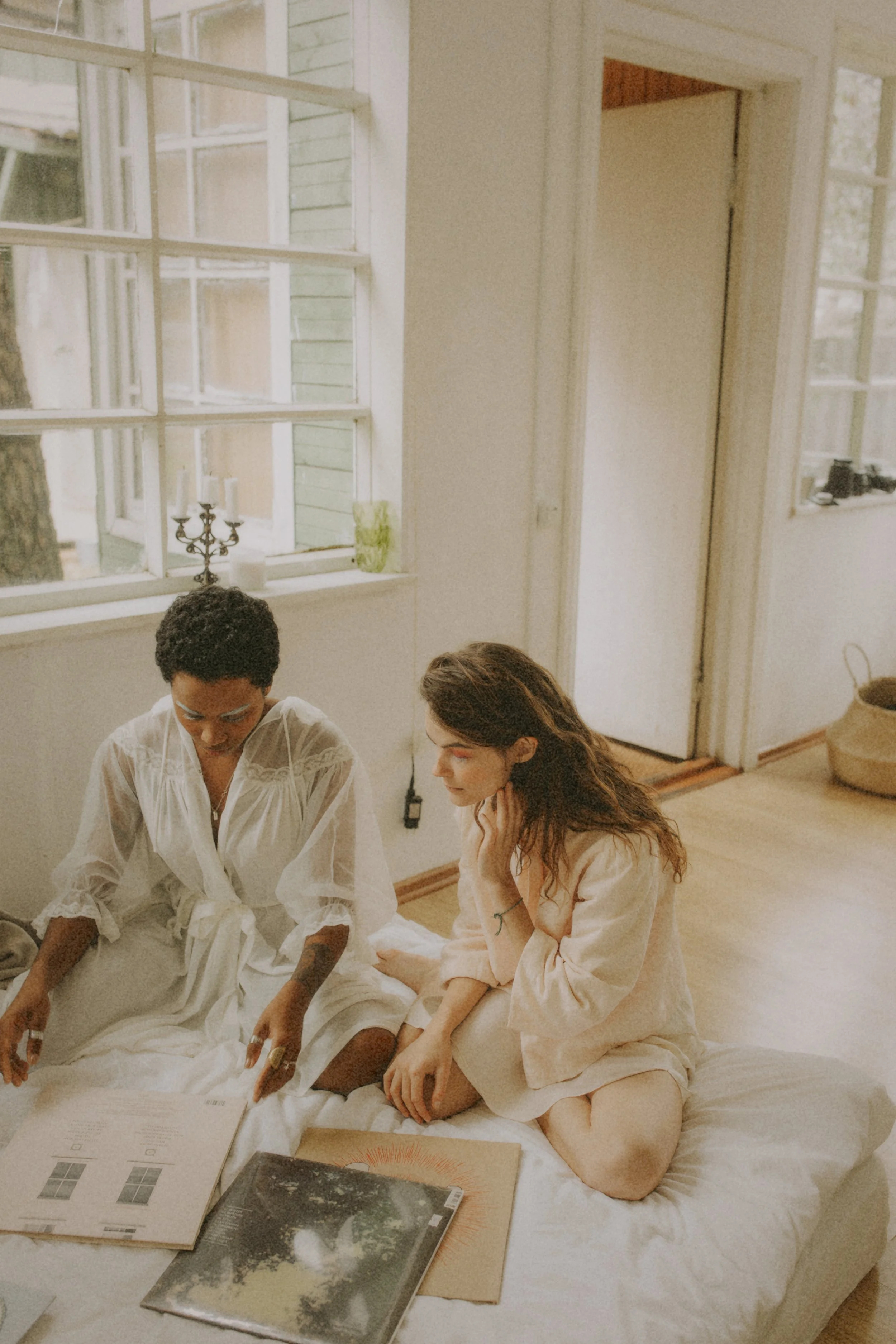 Two women and one man sitting on a bed, reading papers and looking at a book. The woman on the right is sitting on her knees, with her hands on her lap and her chin resting on her hand. The woman on the left is sitting with one knee up and looking at the paper. The man on the left has dark curly hair, wears glasses, and a white long-sleeved blouse. The setting is a cozy room with white walls, windows, and wooden floors. There are magazines, books, and a picture album on the bed.