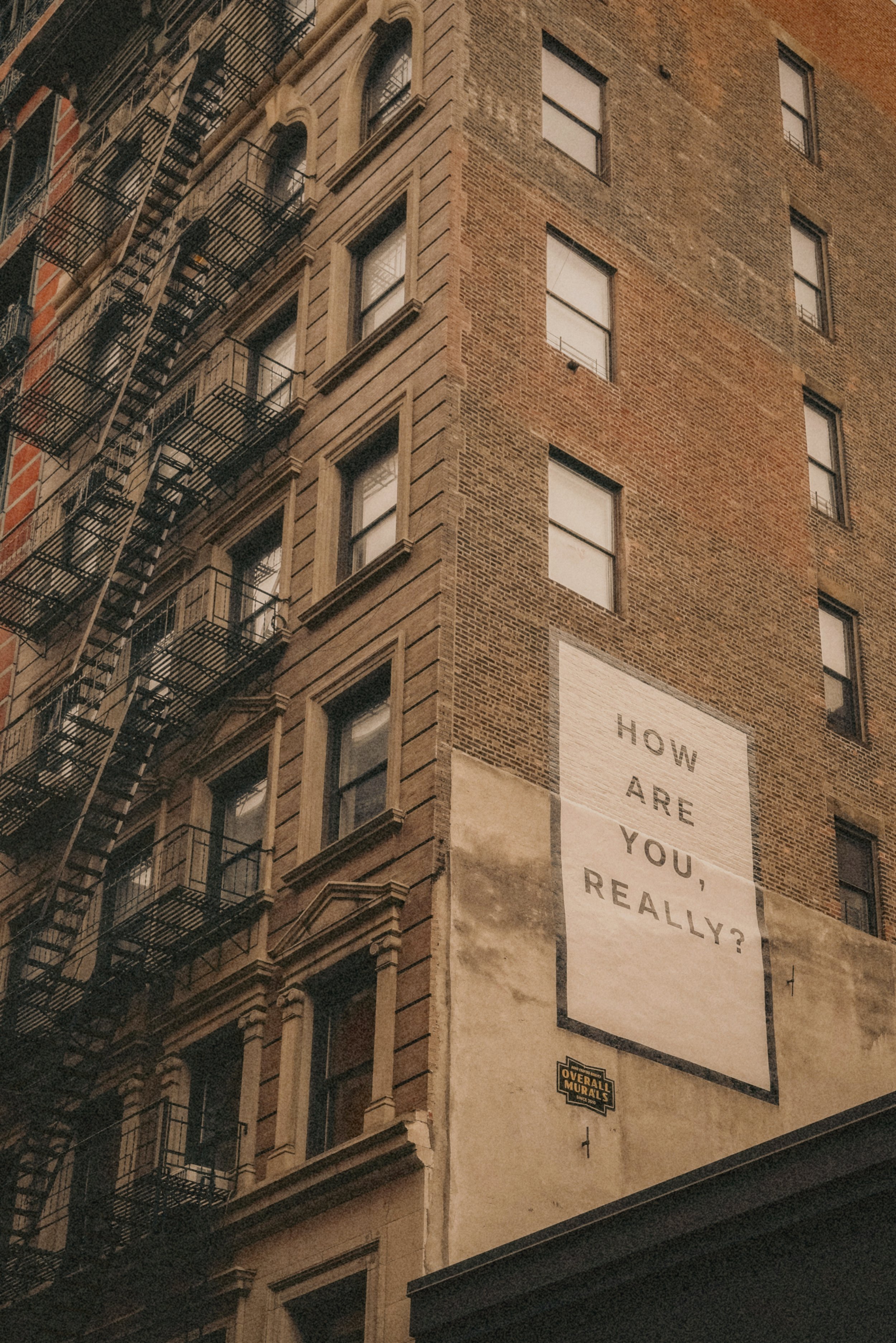 Close-up of a tall brick building with multiple windows and fire escapes, and a large sign that reads, 'How are you, really?'